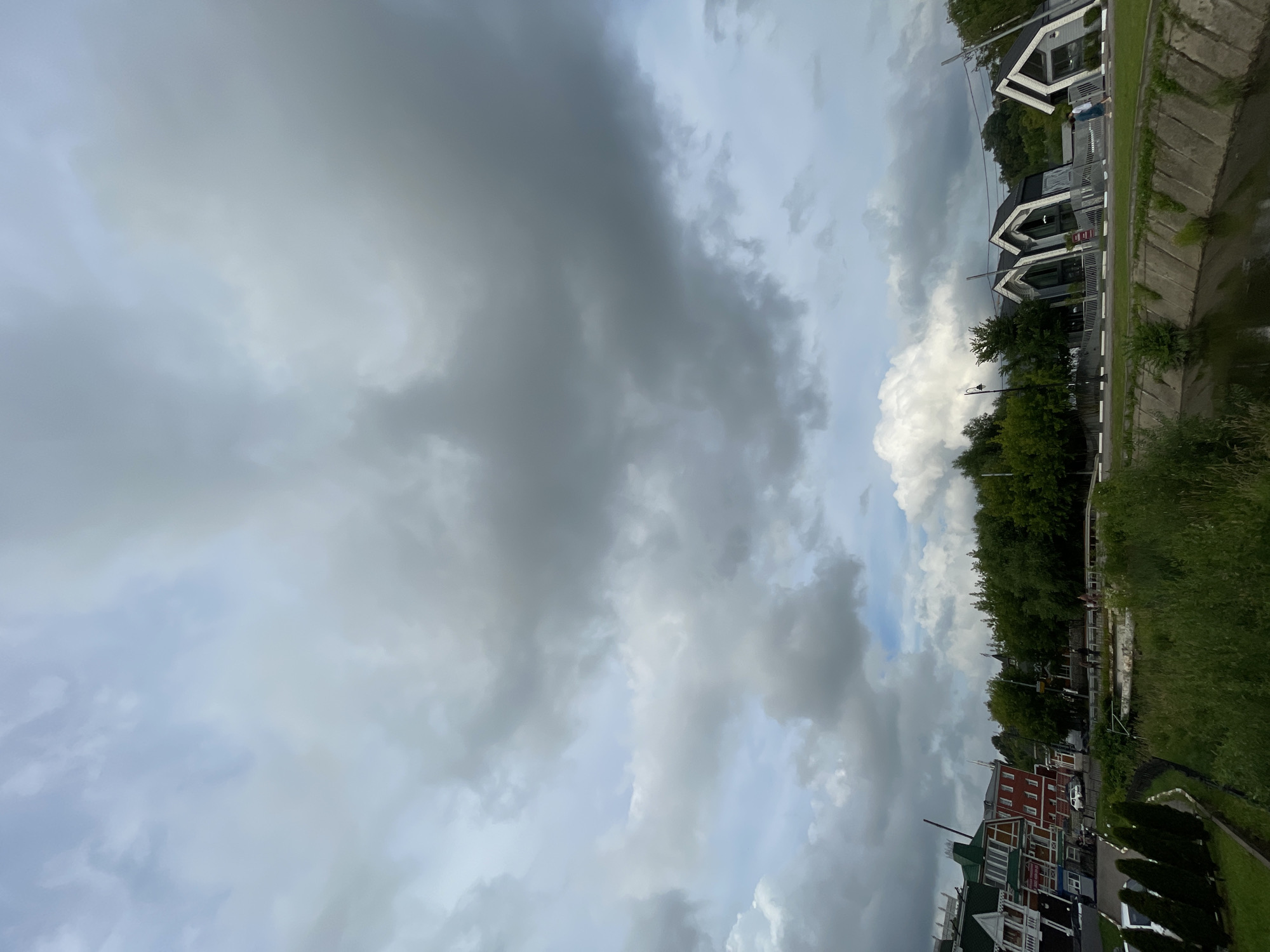 This image, captured in the city of Sergiev Posad, Russia, presents a view of a canal bank lined with green grass and a concrete retaining wall. On the opposite side of the canal, a paved walkway runs alongside a row of distinctive, modern, triangular-roofed buildings. Further back, a line of lush green trees forms a natural barrier. To the left of these modern structures, a section of older, more traditional buildings with red brick and green roofs can be seen. The sky above is heavily overcast with dramatic grey clouds. A few people are visible on the walkway near the modern buildings. The overall atmosphere is that of an urban environment situated next to a natural waterway, under a brooding sky.
