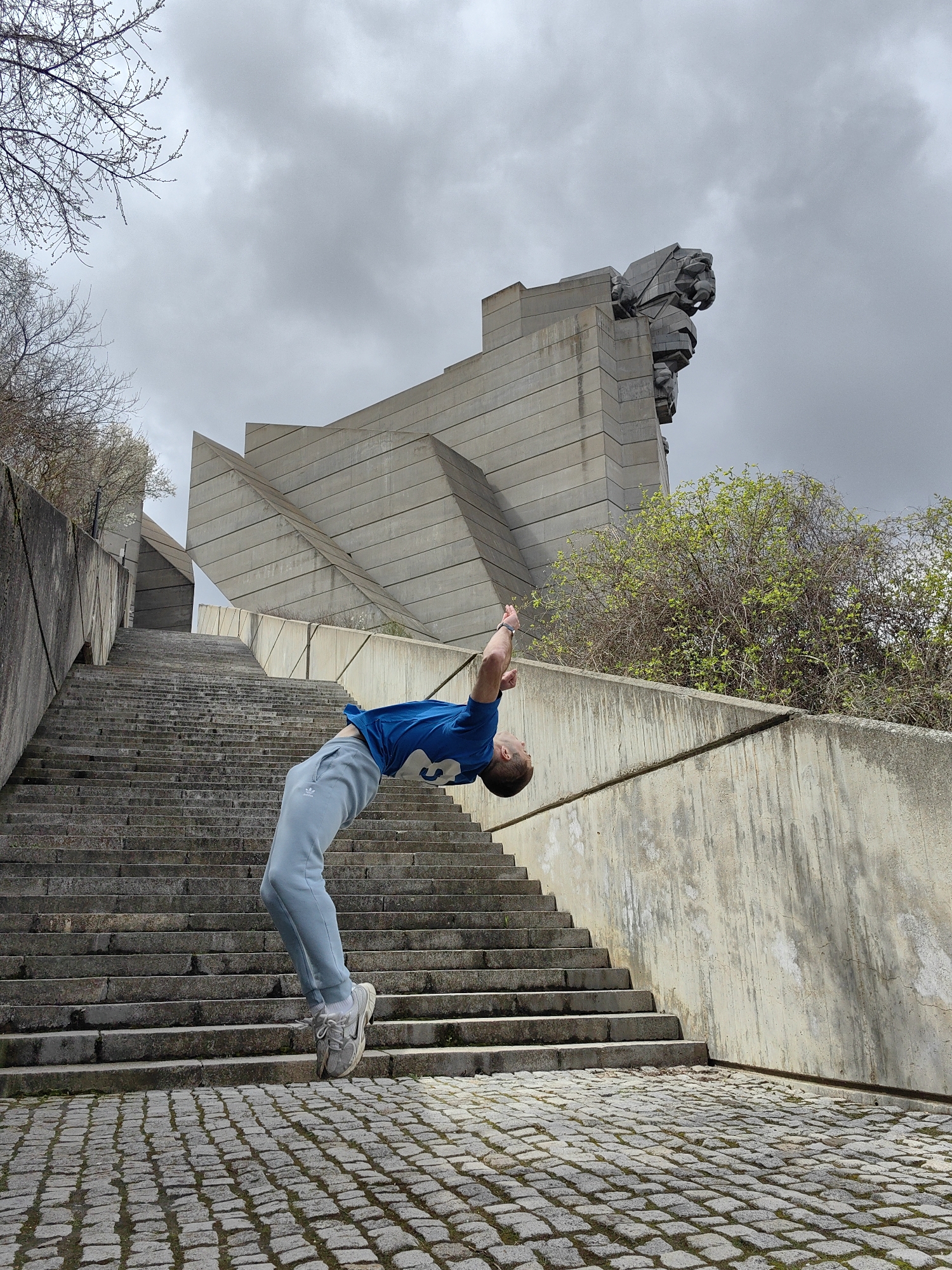 A man in a blue T-shirt and light blue sweatpants is performing a backflip on the steps leading to the Monument to 1300 Years of Bulgaria in Shumen, Bulgaria. The monument is a large, abstract concrete structure featuring stylized figures and geometric forms, including a massive lion head sculpture. The steps are concrete and extend upward towards the monument. The ground at the base of the steps is paved with cobblestones. The sky is overcast. Some bare trees and budding bushes are visible in the background.