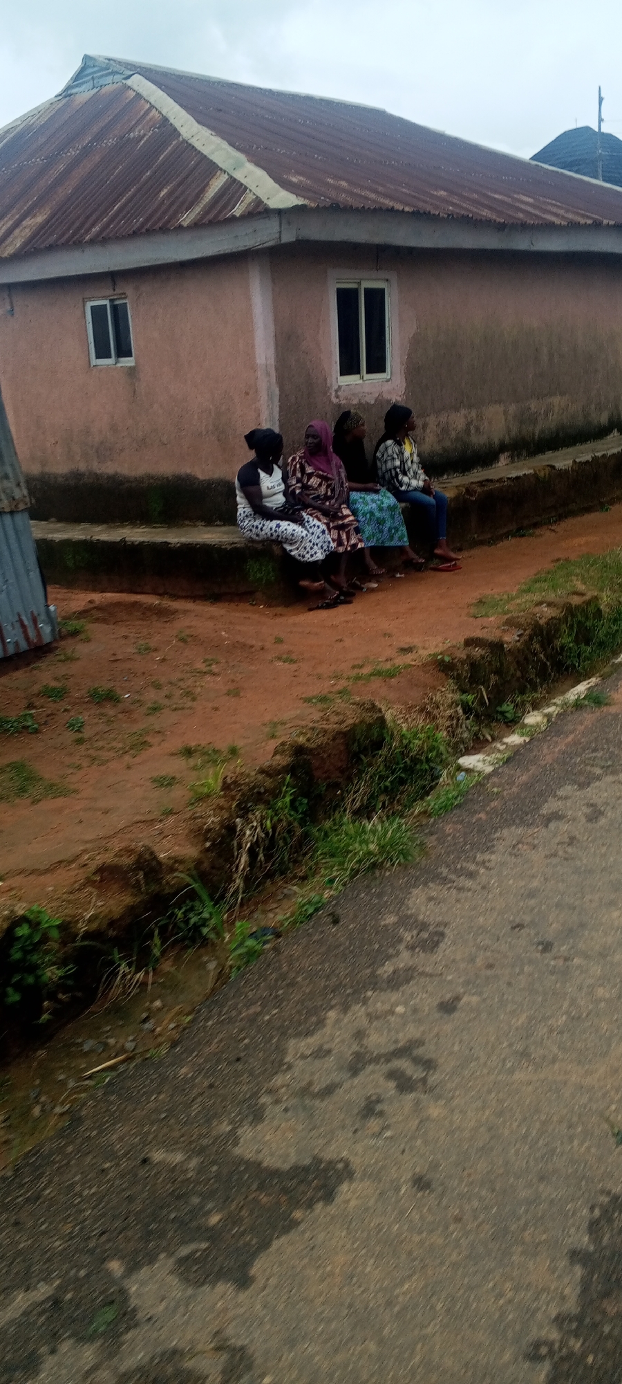 Four women are seated on a concrete ledge in front of a pale pink building with a corrugated iron roof. The building has two windows with white frames. The ground in front of the ledge is a mixture of reddish-brown soil and sparse green grass. To the right of the ledge, there is a dirt ditch with more vegetation. A paved road is visible in the foreground, angled across the image from bottom right to top left. The sky is overcast and a dull grey. The scene appears to be outdoors, in a rural or semi-rural setting, possibly in Bahit, Nigeria, given the architectural style and the surrounding environment. The women are dressed in traditional clothing, with headscarves covering their hair. They are casually seated, some looking towards the building, others looking to the side. One woman wears a white shirt with a graphic design and a patterned skirt, another a patterned dress and pink headscarf, a third a black top and a blue and green patterned skirt, and the fourth a plaid shirt and blue jeans.