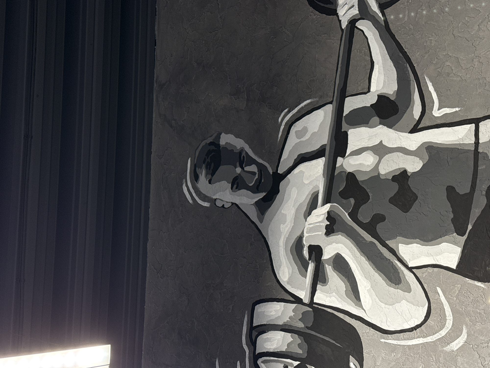 The image depicts a black and white mural painted on a textured wall. The mural features a weightlifter in motion, gripping a barbell loaded with weights. The left side of the image shows a dark, possibly metal, corrugated wall structure. The painting style is graphic, using distinct shades to define the figure and equipment. No other people are visible. The city of Votkinsk, Russia, is not identifiable from the visual information provided in the image.