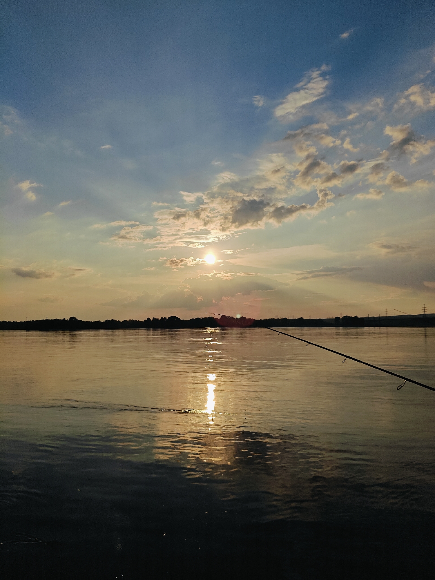 The image depicts a serene sunset over the Kama River in Chaikovsky, Russia. A fishing rod extends into the frame from the right, indicating active fishing. The sun is partially obscured by clouds, casting a warm, golden light across the water and sky. The far bank of the river is silhouetted with trees, and in the distance, power lines are visible. The water is calm, with gentle ripples reflecting the colorful sky and sunlight. The overall atmosphere is peaceful and tranquil, typical of an evening fishing excursion.