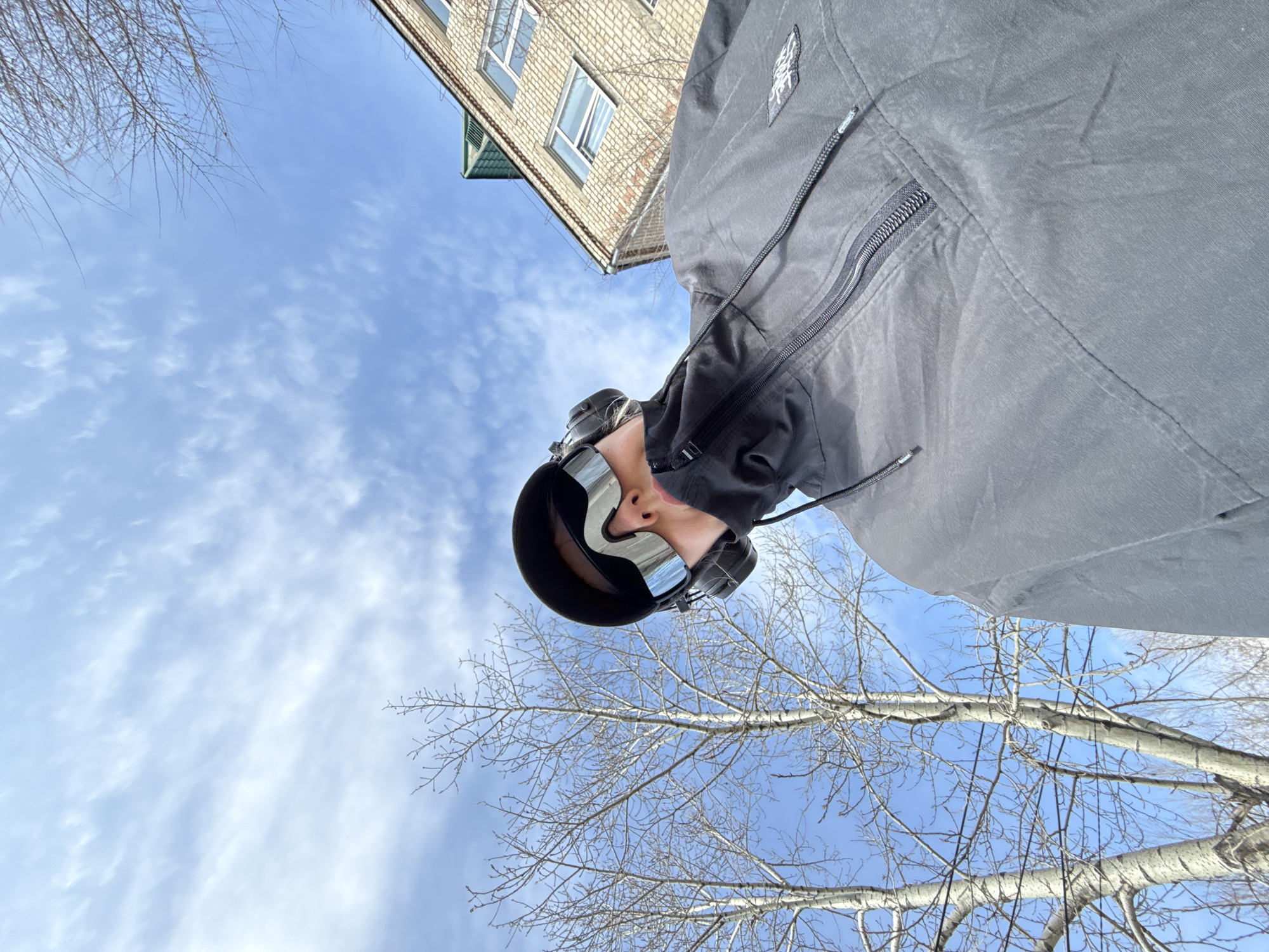 A low-angle, rotated photograph captures a person looking upwards against a bright blue sky with scattered white clouds, likely taken in Borzya, Russia. The image is oriented with the subject's head at the top, seemingly looking down from the sky, while the camera points upwards.
The visible subject is a person wearing large, reflective or mirrored goggles with a dark frame, completely obscuring their eyes and part of their face. They are dressed in a dark grey or black jacket featuring multiple zippers and drawstrings. An indistinct, light-colored logo or text is visible on a patch on the upper left arm of the jacket.
The background consists of a clear blue sky with some wispy clouds. Bare tree branches, characteristic of late autumn, winter, or early spring, frame the left and lower parts of the sky. In the upper right quadrant, a light-colored brick building with several windows and a section of a green roof overhang is visible, suggesting an urban or suburban setting. The bright illumination indicates it is daytime with sunny weather.