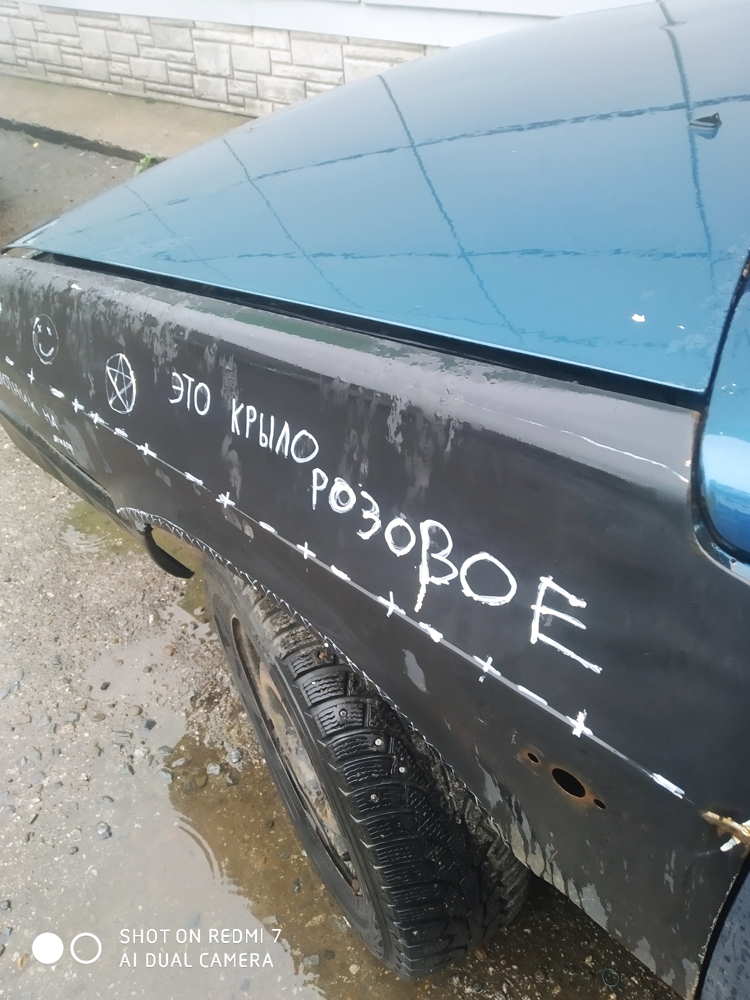 The image depicts the rear passenger side of a blue and black car. The black portion, which appears to be a makeshift or unfinished repair to the rear quarter panel, is marked with white paint. Markings include a smiley face, a pentagram, and Cyrillic text reading "ЭТО КРЫЛО РОЗОВОЕ" (this wing is pink). A series of dashes and crosses are painted along what appears to be a repair line. The car has a tire with visible studs and is parked on a wet, paved surface with visible puddles. The location is identified as Chaikovsky, Russia, based on the device imprint on the image. A wall of light colored stone is visible in the background.