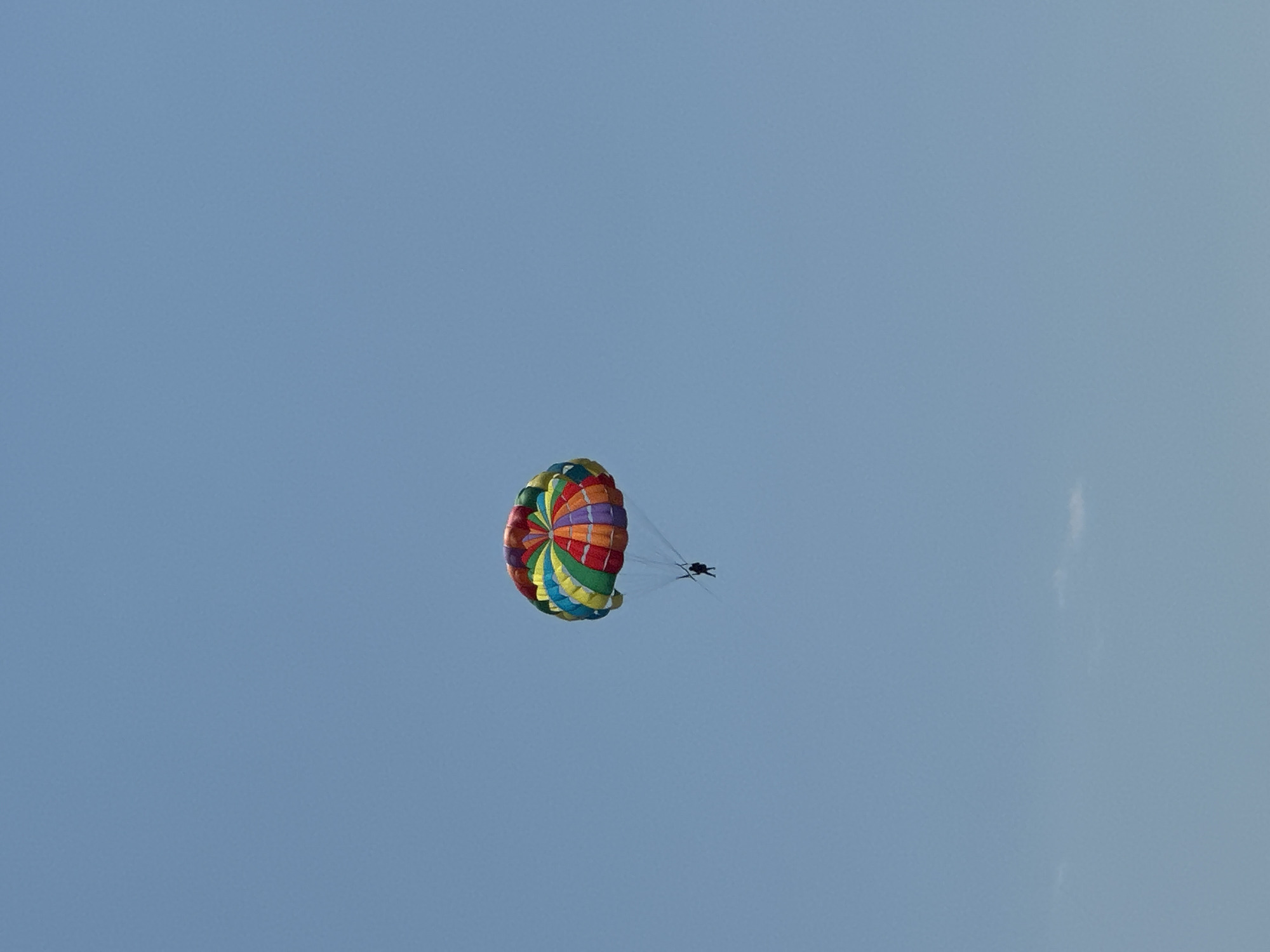 A person is parasailing against a clear blue sky. The parasail is colorful, with panels of red, yellow, green, blue, and purple arranged in a circular pattern. The person is suspended below the parasail by ropes and appears to be a small, dark silhouette. The background is a uniform, cloudless blue sky. There is no indication of the city of Boreti, Montenegro in the image.