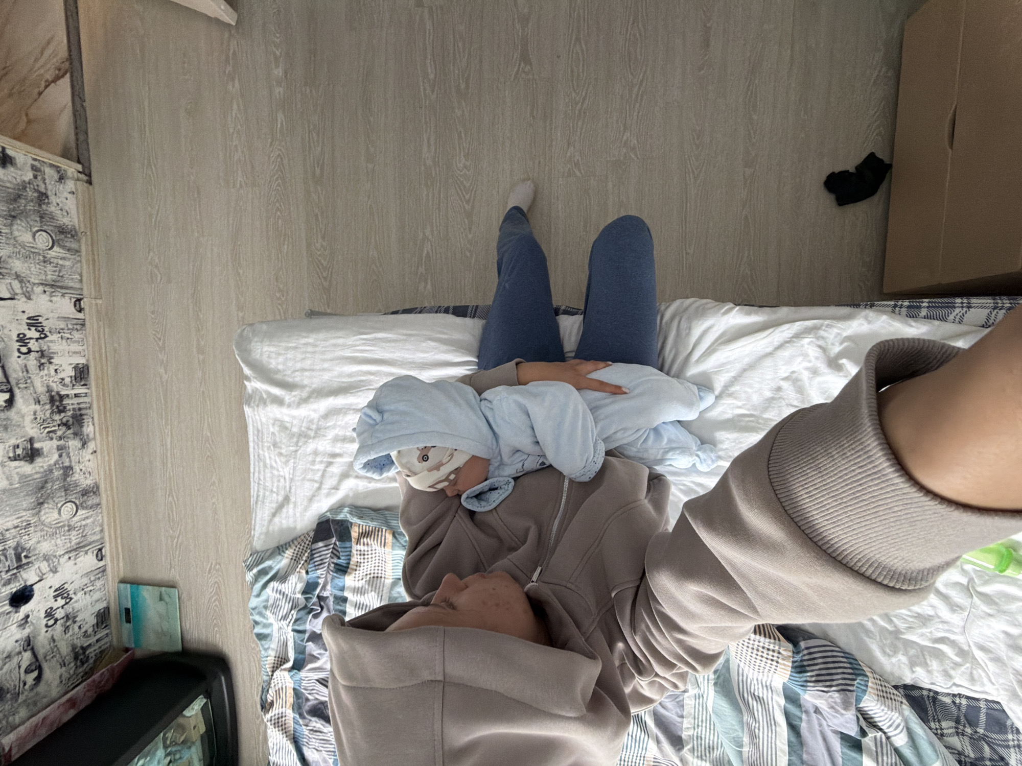A person wearing a beige hoodie and blue leggings is sitting on a bed, holding a baby wrapped in a light blue swaddle. The person is looking down at the baby. The bed is covered with white sheets and a striped comforter. A cardboard box and a pair of black socks are on the floor next to the bed. The floor is made of light wood laminate. A black object, possibly a monitor, is mounted on the wall above the bed. The scene appears to be indoors, likely a bedroom.