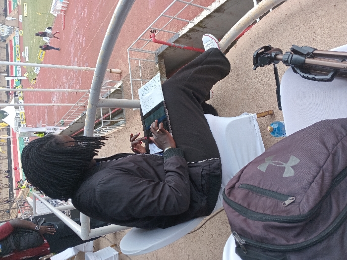 A person with dreadlocks sits at a stadium, working on a tablet. They wear black pants, a black jacket, and white sneakers. A grey Under Armour backpack sits next to them. A tripod and water bottle are also nearby. In the background, a running track is visible, with people running. The scene is captured from a high-angle perspective.