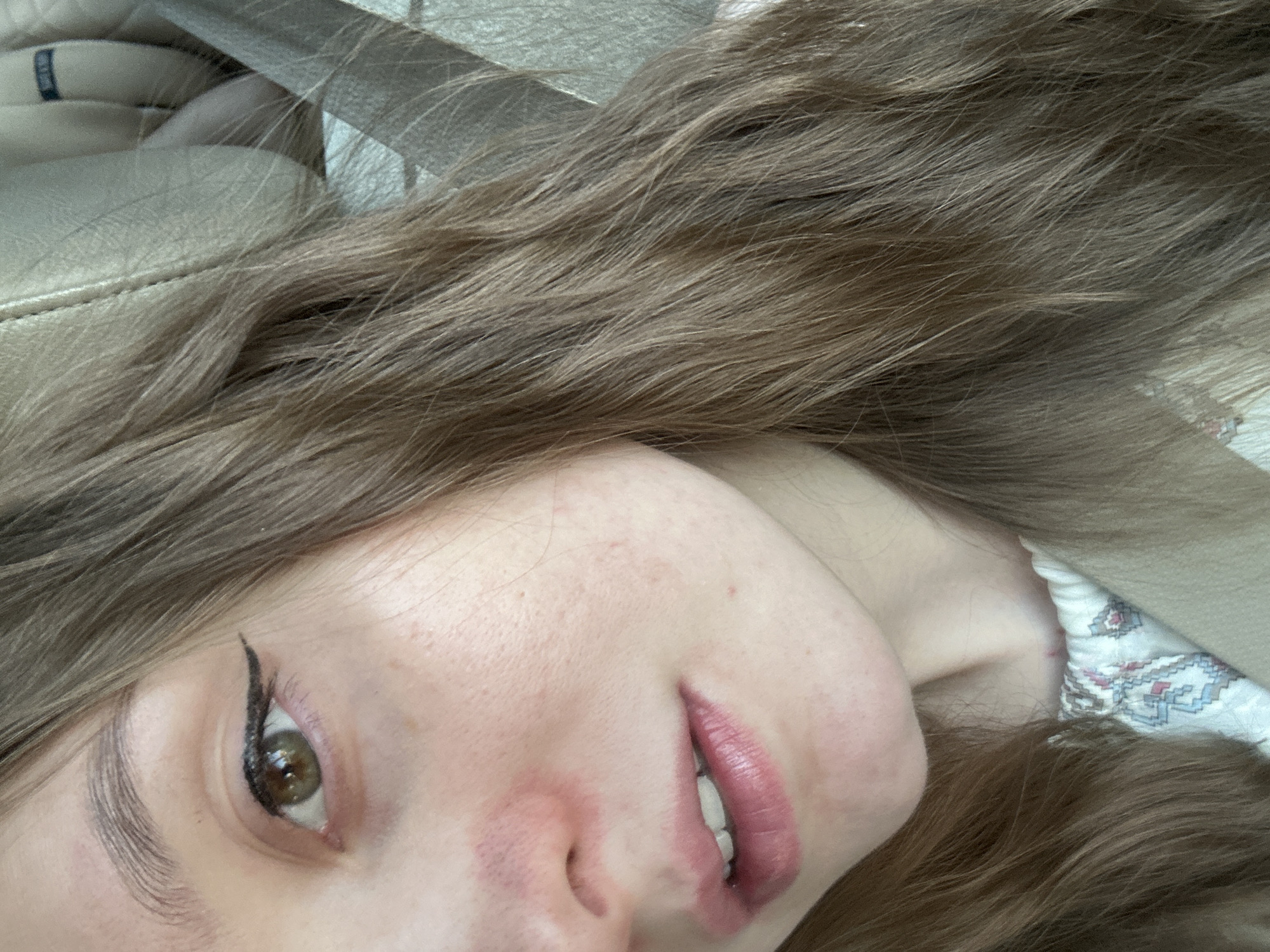 A close-up, angled photograph depicts a woman with light skin and brown hair. She is wearing winged eyeliner and pink lipstick. Parts of her face, including her forehead, nose, and cheeks, are visible. The background includes what appears to be a light-colored leather surface and a patterned fabric. The location in the city of Noviy, Russia is undetermined.