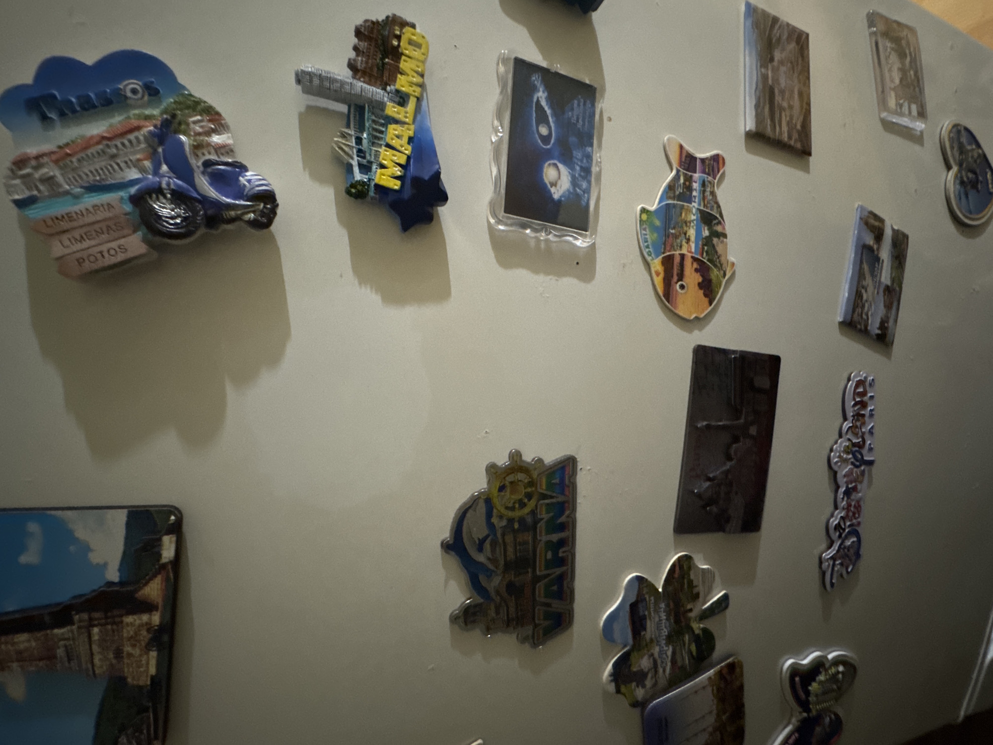 The image shows a collection of refrigerator magnets adhered to a white or light-colored surface. The magnets depict various locations, cities, and themes. Visible magnets include:
* One labeled "Thassos" with images of architecture and a blue scooter.
* One labeled "Malmo" with a bridge image.
* One with a blue and white abstract design in a clear plastic frame.
* A fish-shaped magnet with multiple images.
* One labeled "Varna" with an image including a dolphin and a ship's wheel.
* Others with abstract or landscape imagery.
There is no evidence of Yana, Bulgaria or people in this image. The environment appears to be indoors.