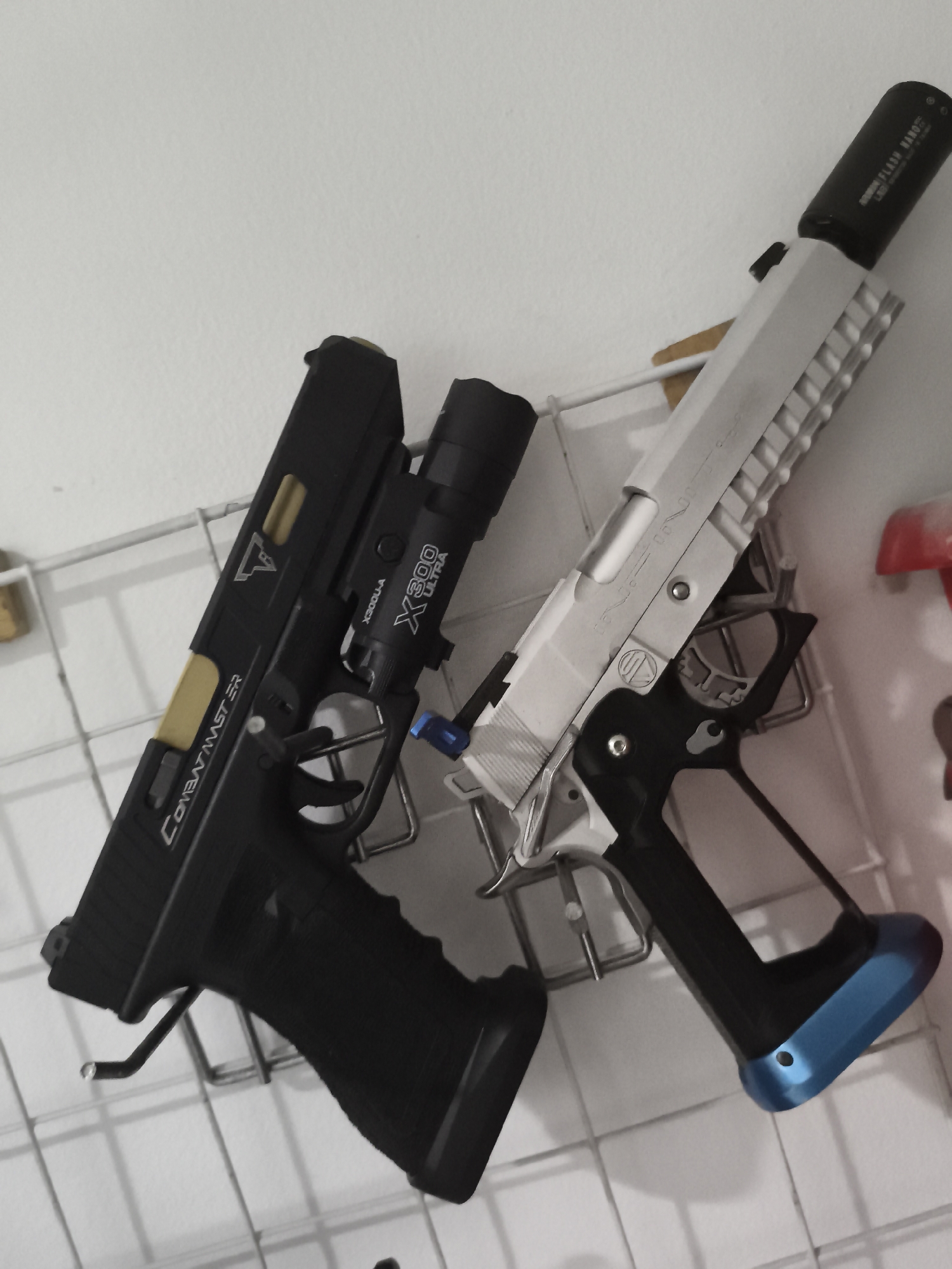 Two airsoft guns are displayed on a white wire grid against a white wall. The gun on the left is black with a gold barrel and a flashlight attached to the front. It has "COMBATMASTER" and "X300-A ULTRA" text visible. The gun on the right is silver with a black grip and a blue base. Both guns are oriented at an angle. The environment appears to be indoors, possibly a display or storage area. The specific location in West Jakarta, Indonesia, cannot be determined from the image.