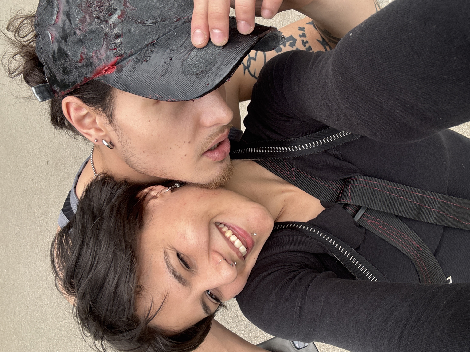 A close-up, high-angle shot features two people embracing. The person on top, a young adult with a dark, stylized baseball cap stained with red markings, has short dark hair pulled back and visible tattoos on their arm. They have fair skin, a light beard, and multiple piercings in their ears. The person below, positioned on their side, has short dark hair and several piercings, including a nose ring and a lip piercing. They are smiling broadly, revealing their teeth. Both are wearing black clothing with straps or harnesses. The setting appears to be outdoors on a paved surface. The city of Sofia, Bulgaria, is not discernible from the provided image.