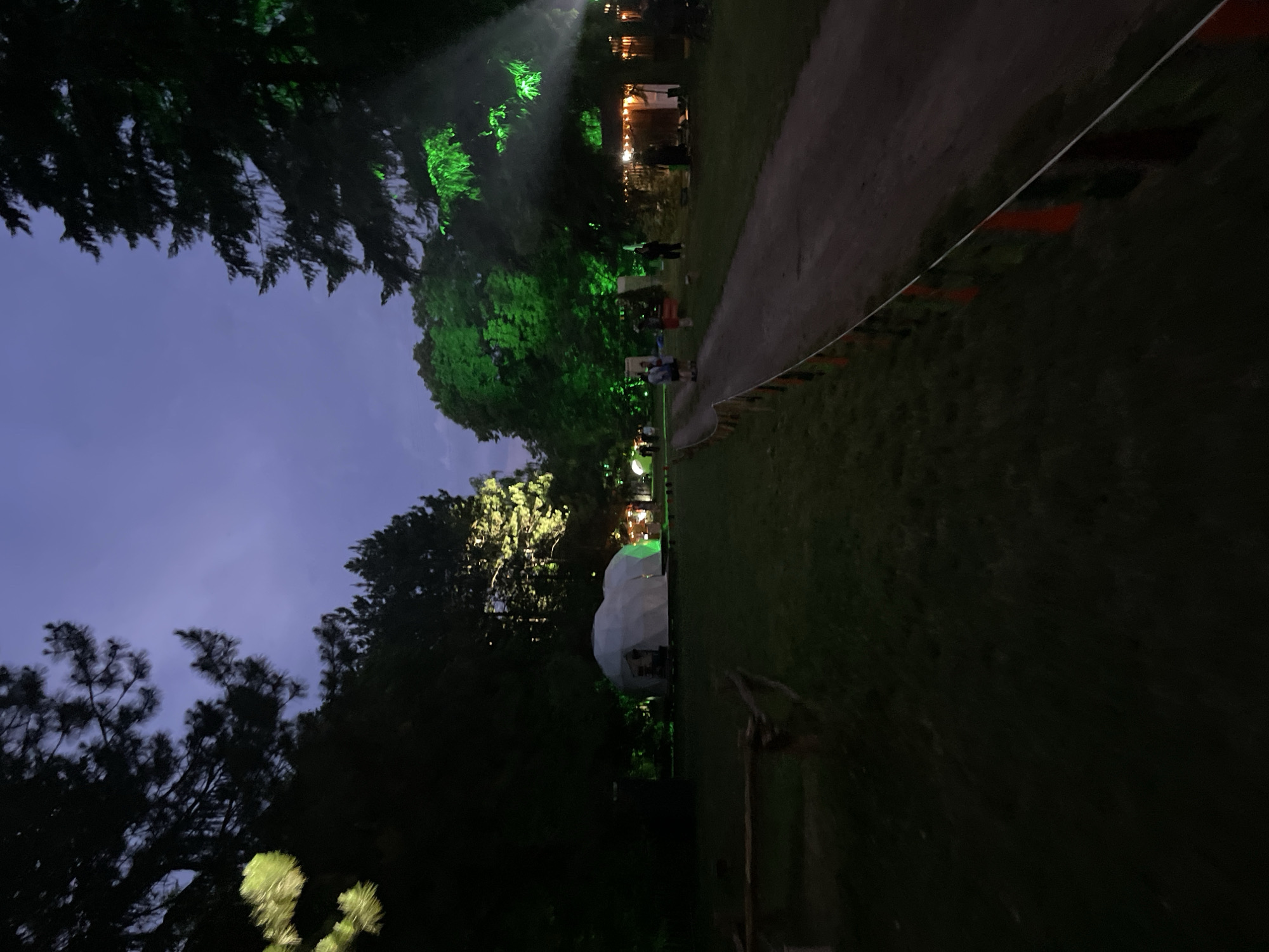 The image captures an outdoor nighttime scene within the Sremski Karlovci Municipality, Serbia. A wide dirt path extends from the foreground into the background, curving slightly to the right. The sky above is a dark blue-purple, indicative of late twilight or early night.
Dense, dark trees border the scene on both sides, with some branches silhouetted against the sky and others vividly illuminated by artificial lights. On the left side of the frame, a large, white geodesic dome or spherical tent structure is visible, with its base bathed in green light. Further into the background, a series of smaller structures, possibly stalls or temporary buildings, are illuminated by warm, yellow string lights. The foliage of several large trees in the midground and right side of the image is intensely illuminated by bright green spotlights, creating distinct glowing effects and visible light beams.
Along the right edge of the dirt path in the foreground, a low demarcation line is marked by a white rope strung with small, alternating red and dark-colored triangular flags. Multiple individuals are present throughout the scene. Two figures are visible on the dirt path in the midground, one appearing to wear a light-colored top. Additional people are discernible near the illuminated structures in the background. The ground consists of dark grass flanking the central dirt path. The scene is dominated by artificial lighting, which casts vibrant green hues on the vegetation and structures, contrasting with the overall darkness and the warm tones of scattered string lights.