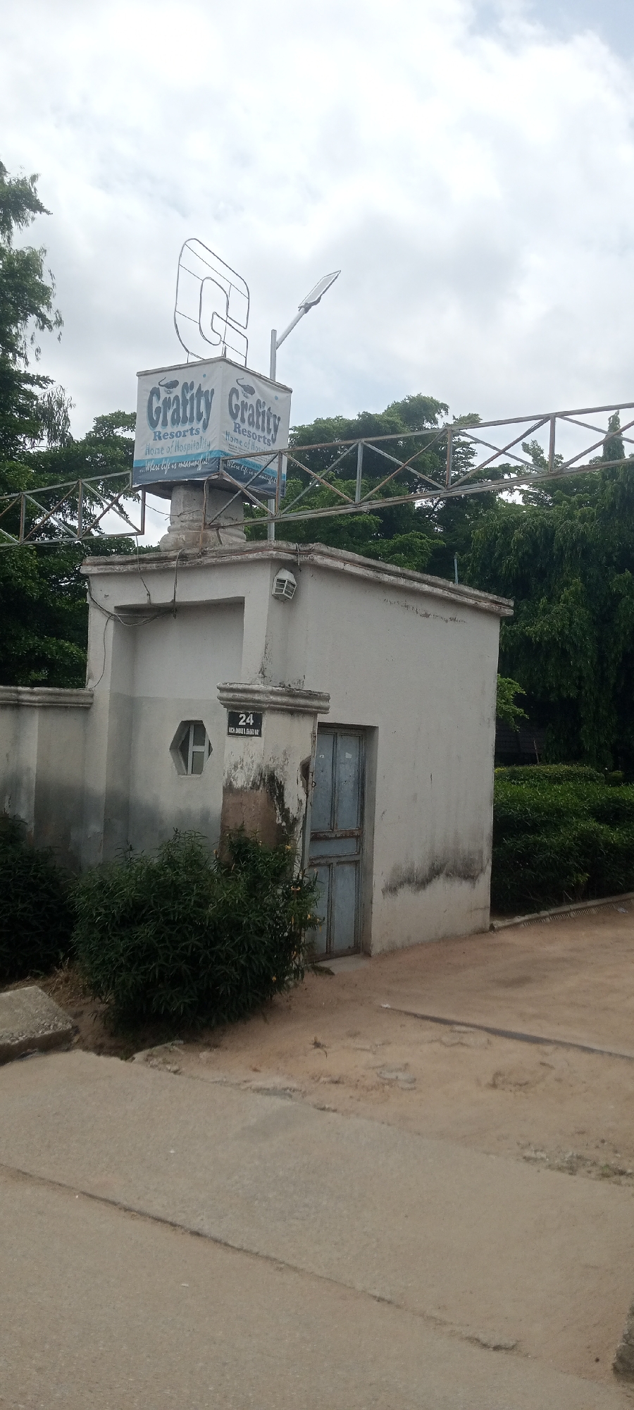 The image shows the exterior of a building with a sign that reads "Grafity Resorts, home of hospitality. Where life is meaningful." The building is painted white and has a blue door. There is a small, hexagonal window on the left side of the building. A sign with the number "24" is affixed to a pillar near the door. The building appears to be located in a somewhat overgrown area, with bushes and trees surrounding it. The sky is cloudy. The overall impression is of a modest resort facility. The location in Jalingo, Nigeria, is not explicitly visible in the image but can be inferred from external context.
