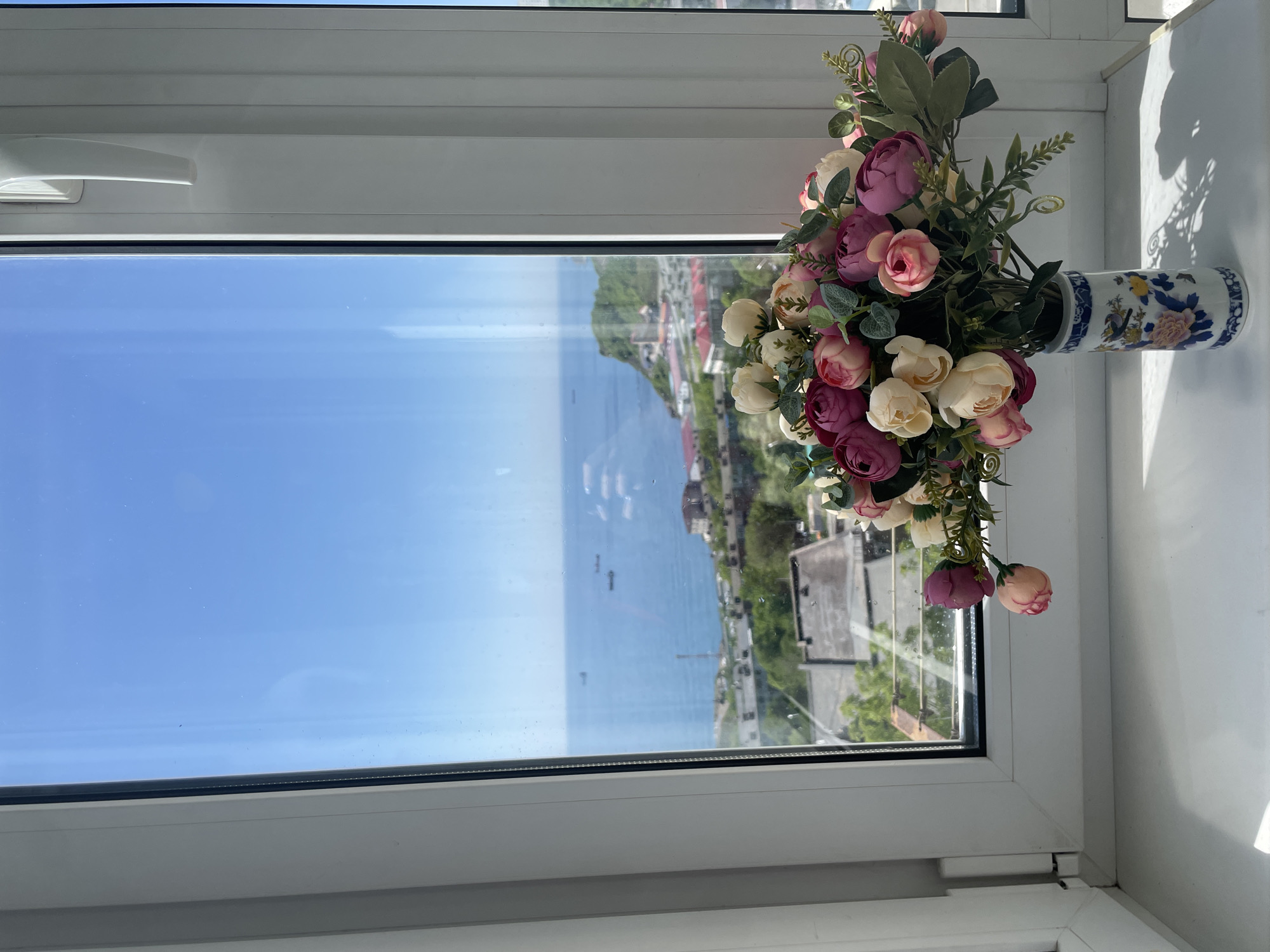 A high-angle, brightly lit shot depicts a window sill with a decorative vase and a view outside to Vladivostok, Russia.
The vase, cylindrical with a narrow neck, sits horizontally on the white window sill. It features a blue and white pattern, with glimpses of yellow and purple floral designs. The vase contains an arrangement of artificial flowers in various colors: white, pink, and purple. Green foliage intermingles with the flowers.
The window provides a view of the city. The sky is clear and blue. Below, water extends towards a landmass, suggesting a coastal area. Buildings and vegetation are visible along the coast, reflecting a mix of urban and natural environments typical of Vladivostok's landscape. Shadows from the objects on the window sill are cast onto the white surface. The window frame itself is white.
