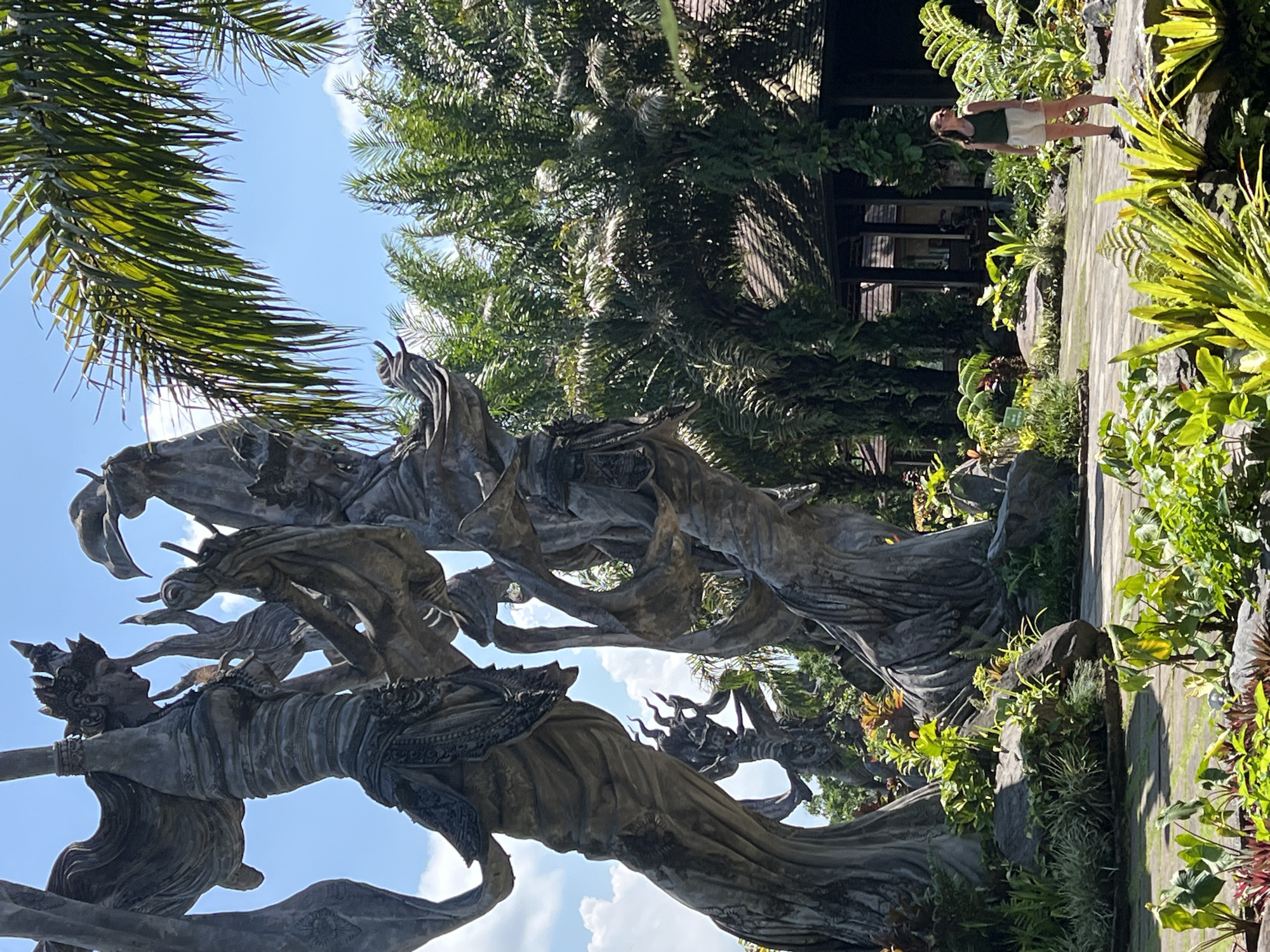 The image depicts a sculpture in a garden setting in Kedewatan, Indonesia. The sculpture appears to be made of gray stone and depicts figures with intricate details. Lush greenery surrounds the sculpture, including palm trees and other tropical plants. A woman stands on a paved path that runs through the garden. The sky is blue with scattered clouds. A building with a dark roof is visible in the background.