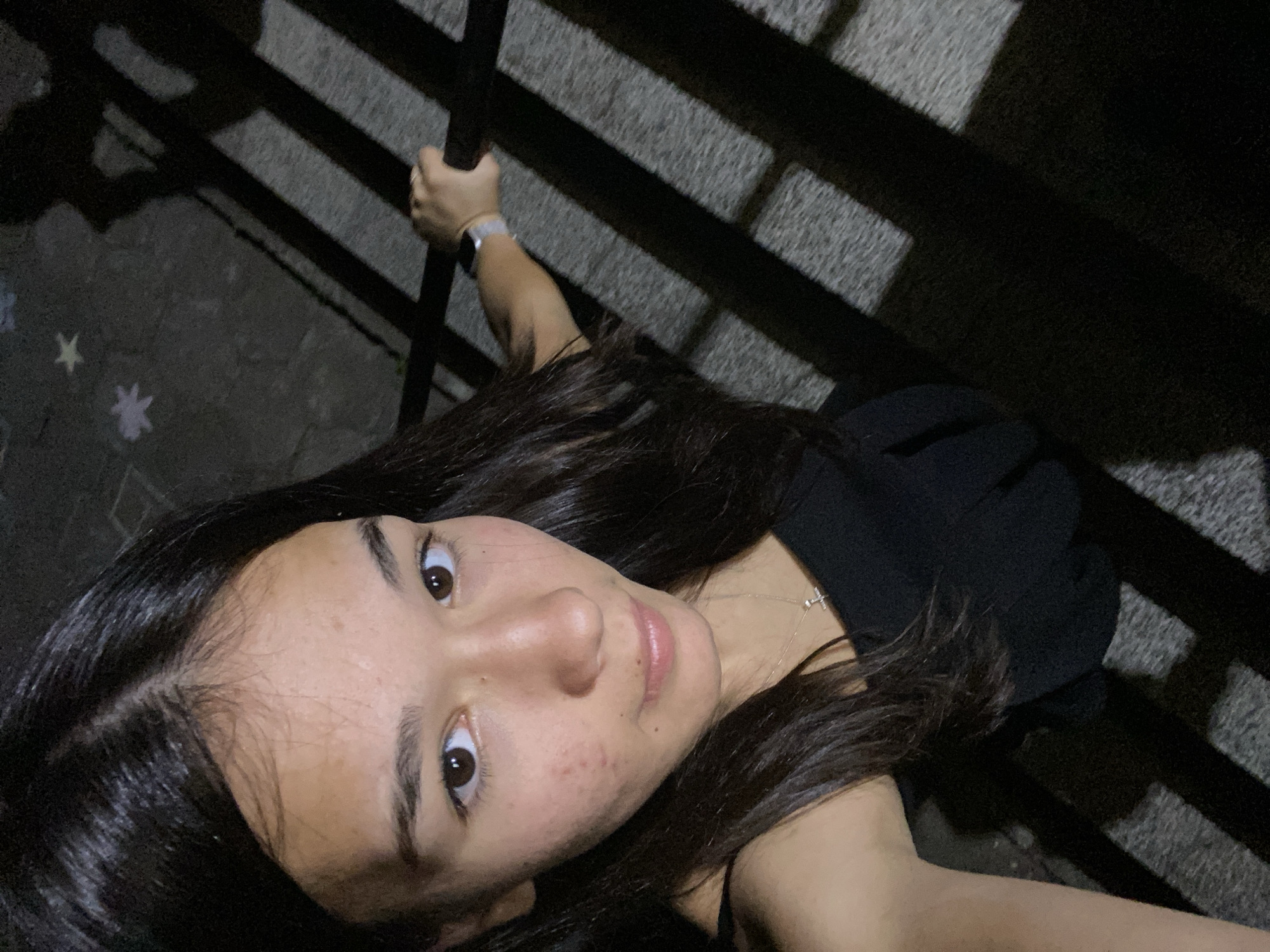 In this high-angle, self-portrait, a young woman with dark hair and brown eyes is pictured at night. She is wearing a black sleeveless top and a delicate silver necklace. Her right hand rests on a railing, and she is wearing a smartwatch on her left wrist. The background consists of a textured ground illuminated by harsh, striped shadows, suggesting a dimly lit outdoor urban setting. There are two small, light-colored star-shaped objects visible on the ground near the upper left of the image. No specific location in Hirevo, Bulgaria, can be determined from the image.