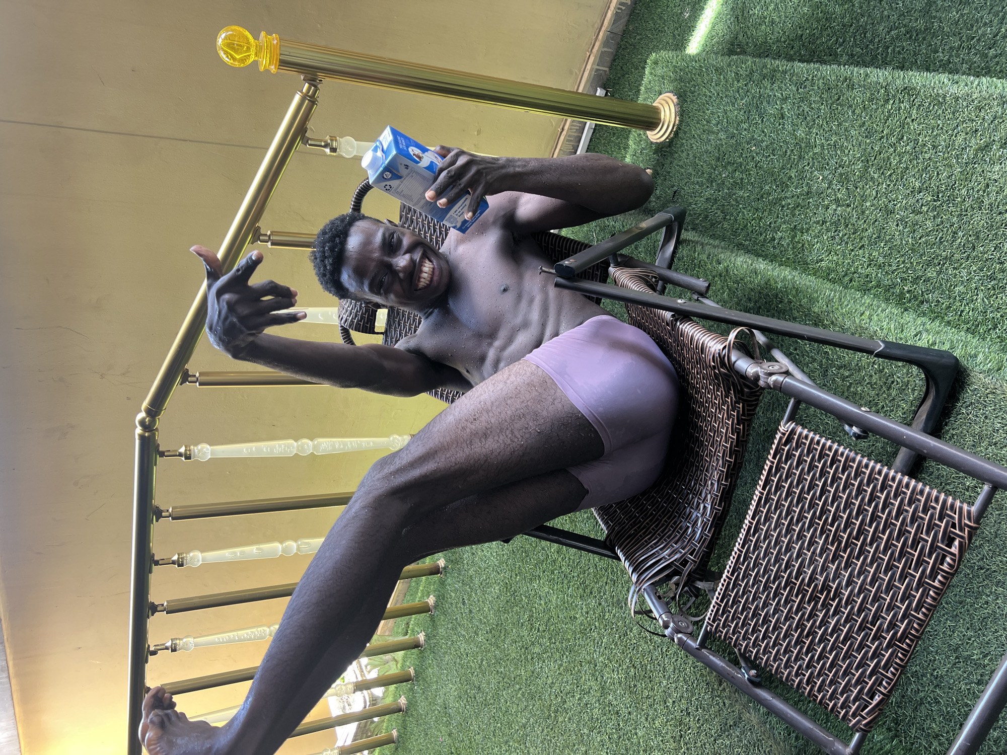 A man is sitting in a wicker chair outdoors. He is holding a blue carton and smiling. He is wearing light purple underwear. There is a gold metal railing to his left and artificial green grass is on the ground behind him. The location is unknown.