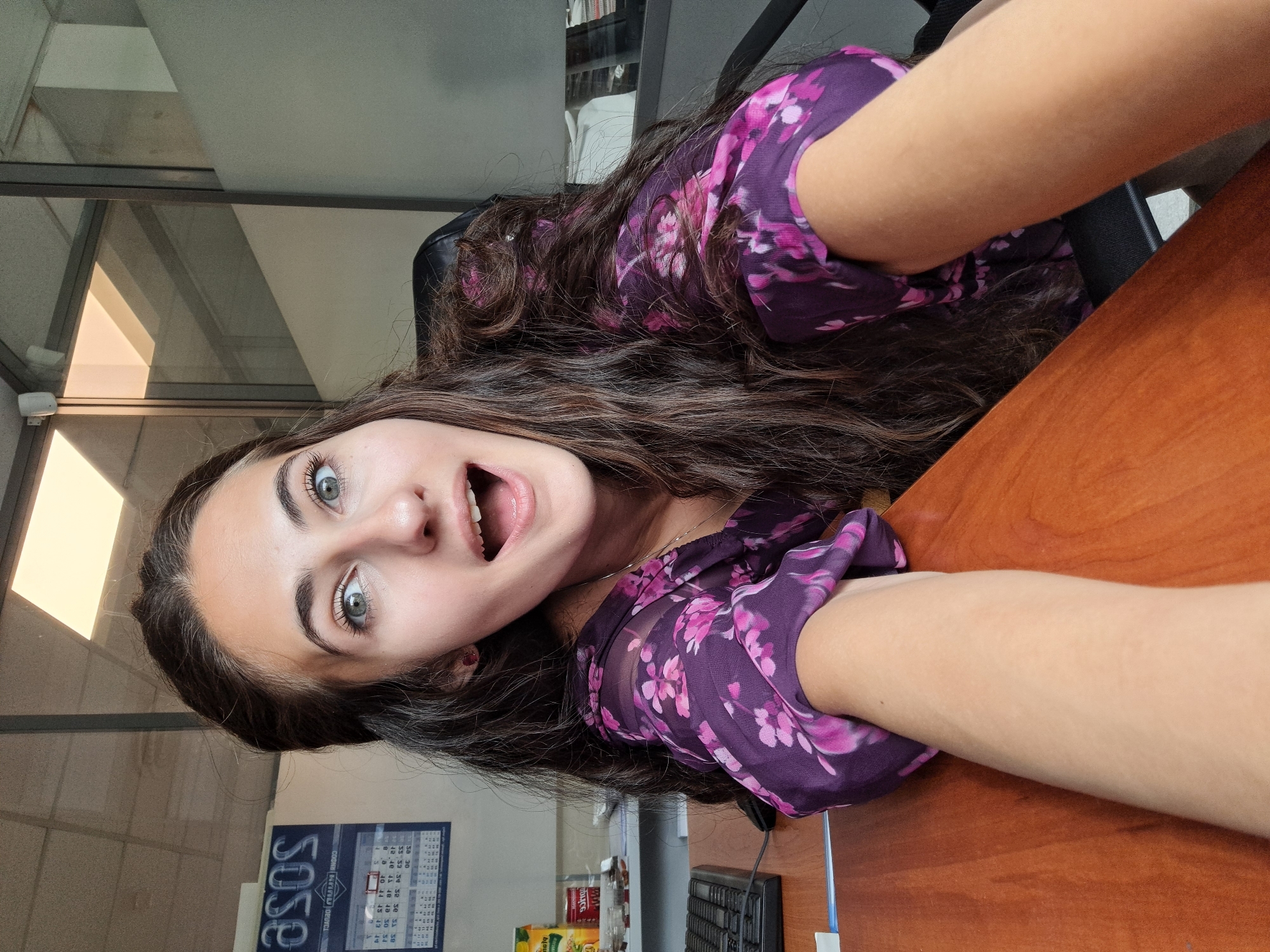 A young woman with long, dark brown, wavy hair and green eyes is captured in a close-up selfie, taken indoors within what appears to be an office or commercial setting. She looks directly at the camera with an expressive, surprised, or excited facial expression, her mouth open and eyes wide. Her arms are extended, holding the camera to take the picture.
She is wearing a sheer, dark purple blouse with a distinct pink floral pattern, and a thin silver necklace is visible around her neck. Her skin is fair.
The background shows elements of an indoor work environment, including a wooden desk in the foreground on which a black computer keyboard and various other items, such as a yellow packet and possibly food containers, are placed. A black office chair is partially visible behind her. In the background, there are glass partitions or windows, reflecting interior ceiling lights. A blue sign or poster is visible on a wall or partition, displaying the reversed text "SOSO", along with a calendar showing numbers. The image is rotated sideways, implying it was taken in a portrait orientation.
