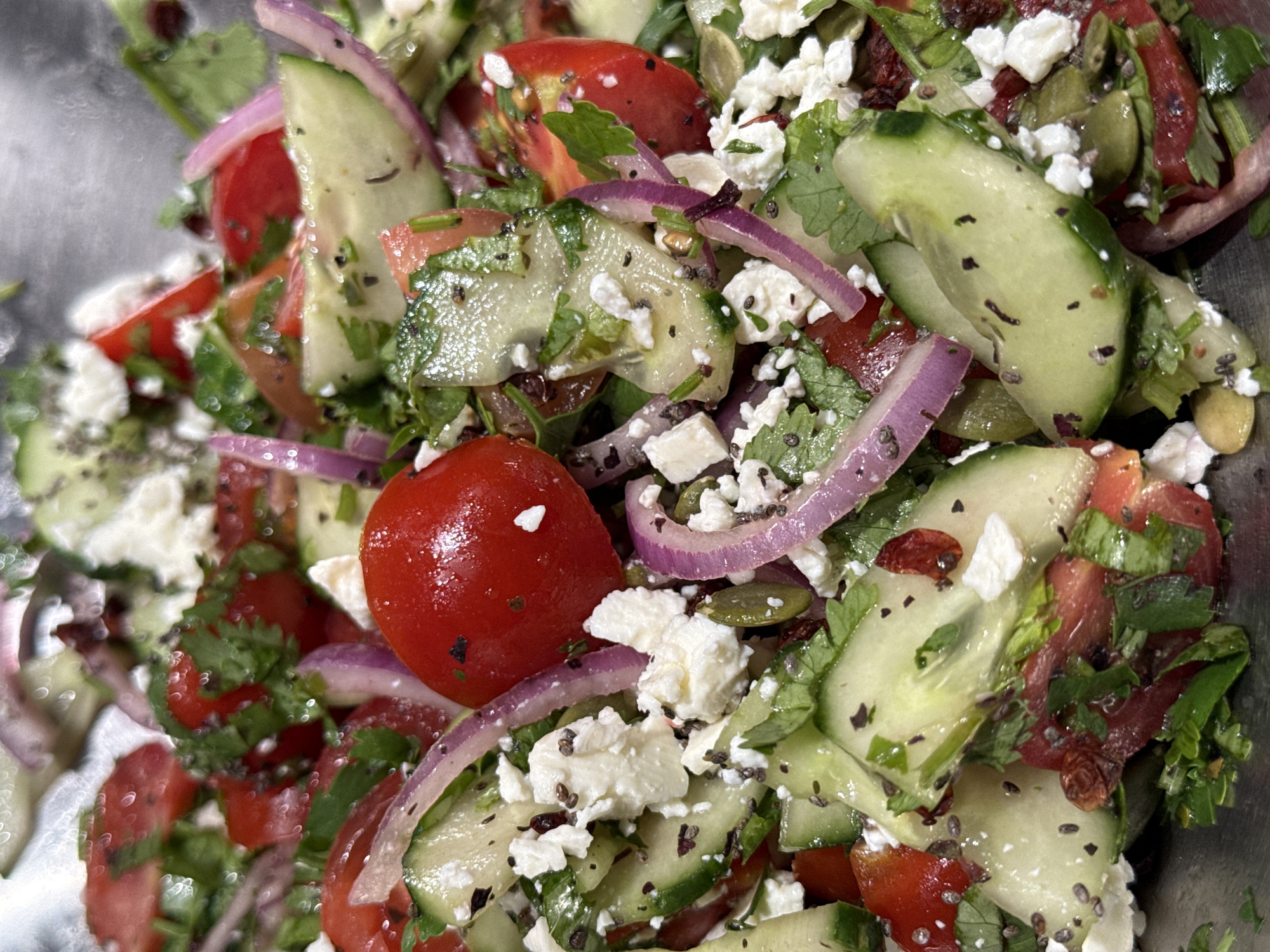 The scene depicts a mixed salad in Yerevan, Armenia. Contained within a metallic vessel, the composition includes sliced cucumbers, halved red cherry tomatoes, thin red onion slivers, and crumbled white cheese. Chopped green herbs, consistent with cilantro or parsley, are interspersed throughout. Small black seeds, resembling chia seeds, and green pumpkin seeds (pepitas) are distributed as garnish. Vegetable surfaces exhibit a moist sheen, indicative of a liquid dressing. No human subjects are present, and no actions or interactions are depicted.