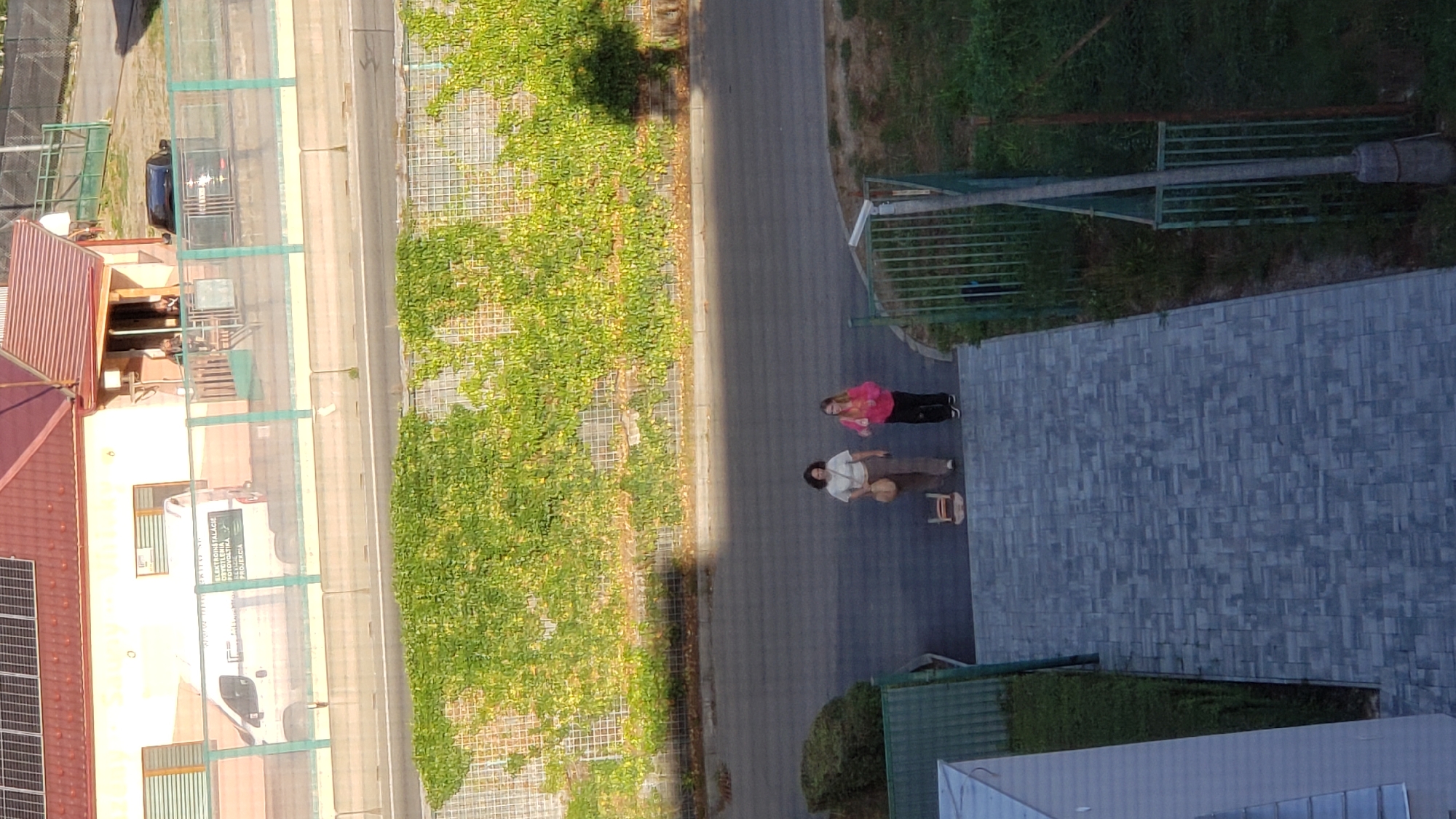 In Prešov, Slovakia, two women are walking on a paved path adjacent to a road. The woman on the left wears a light-colored shirt and pants, carrying a straw bag. The woman on the right wears a pink hooded jacket and black pants. A small wooden stool is placed on the path between them. The scene takes place outdoors, with a building featuring solar panels visible in the background, along with a white van parked near the building. Green fencing separates the building area from the road. Lush green vegetation covers a wall in the middle ground. The overall environment suggests an urban or suburban setting.