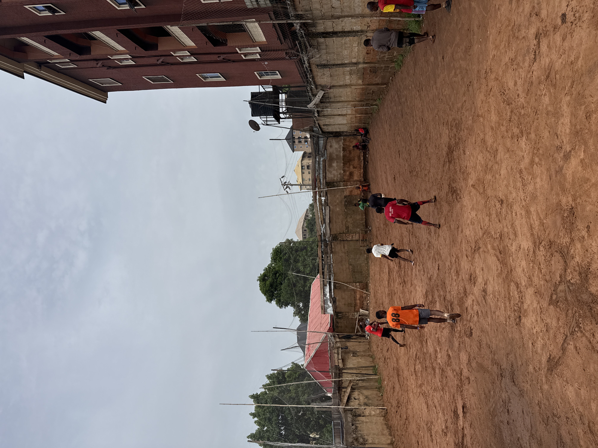 A medium shot captures a soccer match in Nkpor, Nigeria. The players, attired in sports wear, engage in the game on a dirt field surrounded by buildings and fences. Overhead, a cloudy sky indicates possible rain. The surroundings feature both residential and commercial structures, with a water tower. Power lines are visible.