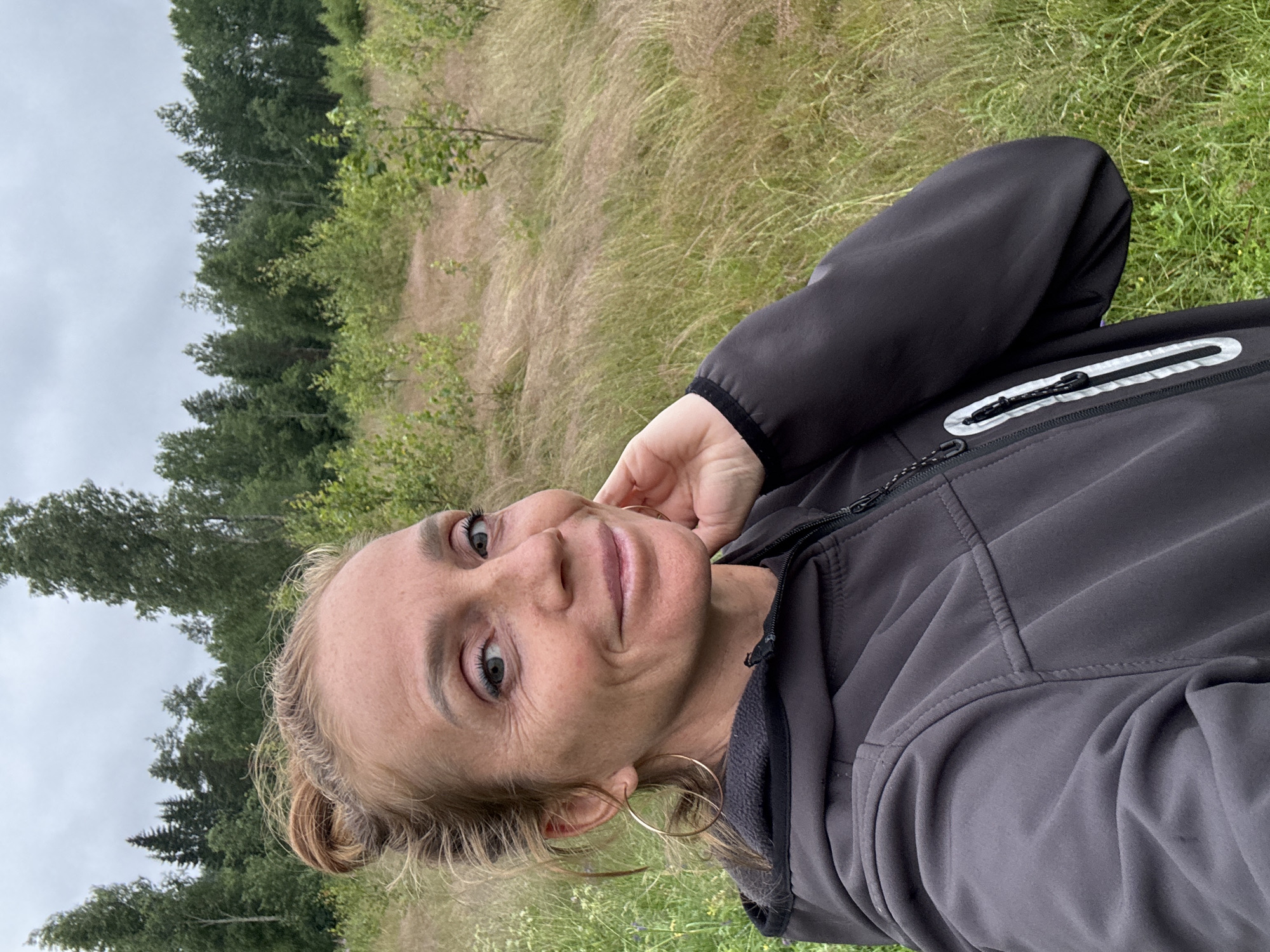 A woman with blonde hair pulled back is taking a selfie in an outdoor setting. She is wearing a dark grey jacket with a zipper and reflective detailing. Her left hand is resting on her chin. She is wearing large hoop earrings. The background consists of green grass, tall dry grass, and a line of trees. The sky is overcast, suggesting cloudy weather. The image appears to be taken in daylight. The location cues, such as the type of vegetation, are consistent with a natural, potentially rural or wilderness area. The woman's expression is relaxed and friendly, with a slight smile.
