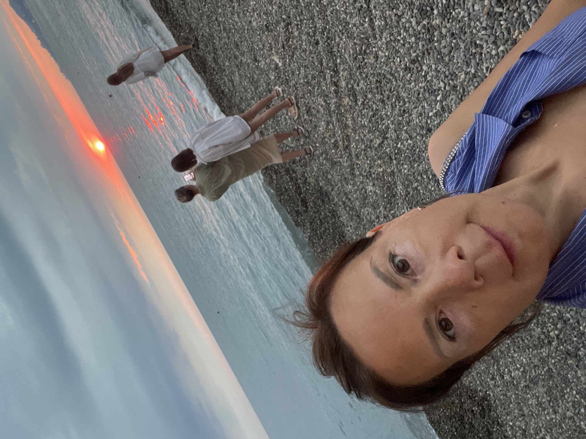 In Sochi, Russia, a selfie captures a woman in the foreground, her face close to the camera, with a pebbled beach and the Black Sea stretching behind her. The sky is overcast, with a vibrant orange sunset breaking through the clouds, casting a warm glow on the water. Three other figures are visible further down the beach. Two of them, a person in a light green top and skirt and another in a white shirt, are standing close together, with the person in green holding up a phone, seemingly taking a photo. A third person in a white dress stands further away, looking out at the sea. The waves are gently lapping at the shore.