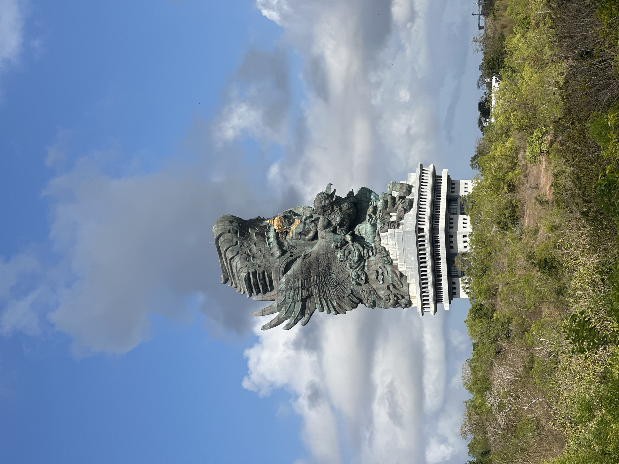 The image shows a tall, intricately designed statue of Garuda, the mythical bird-like creature from Hindu mythology, standing atop a multi-story building. The statue is made of a dark, metallic material and features large, spread wings, a human-like torso with ornate jewelry and a crown, and a bird's head. The building beneath the statue is modern, with a light-colored facade and a series of windows. The statue and building are situated on a hill covered with dense green and brown vegetation. In the background, the sky is blue with scattered white and gray clouds, and a few distant birds are visible. The scene is located in Kuta Selatan, Indonesia, identified as the Garuda Wisnu Kencana Cultural Park. There are no people or animals visible in the immediate foreground or on the statue itself. The overall environment is outdoor and appears to be a tourist attraction or cultural landmark.