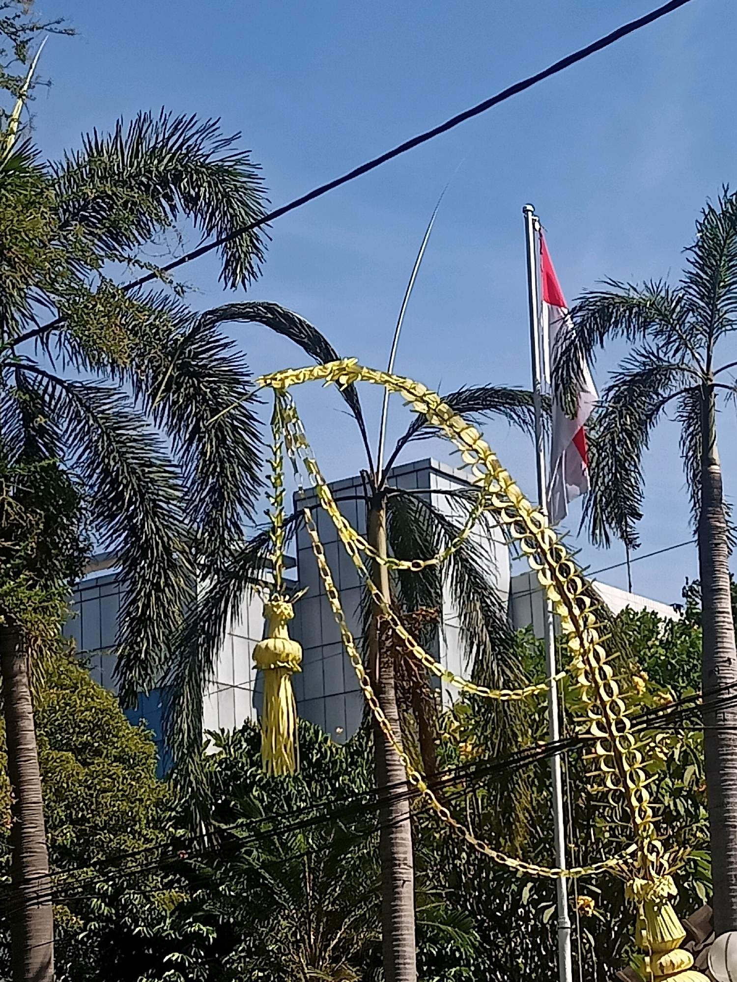 The image shows a street scene in Surakarta, Indonesia, with palm trees and lush green foliage in the foreground and midground. A modern building with glass windows is visible in the background. A flagpole with the Indonesian flag, red and white, stands prominently against a clear blue sky. Decorative yellow chains and a yellow tassel-like ornament are suspended between the palm trees, suggesting a festive occasion or decoration. The scene is illuminated by bright sunlight.
