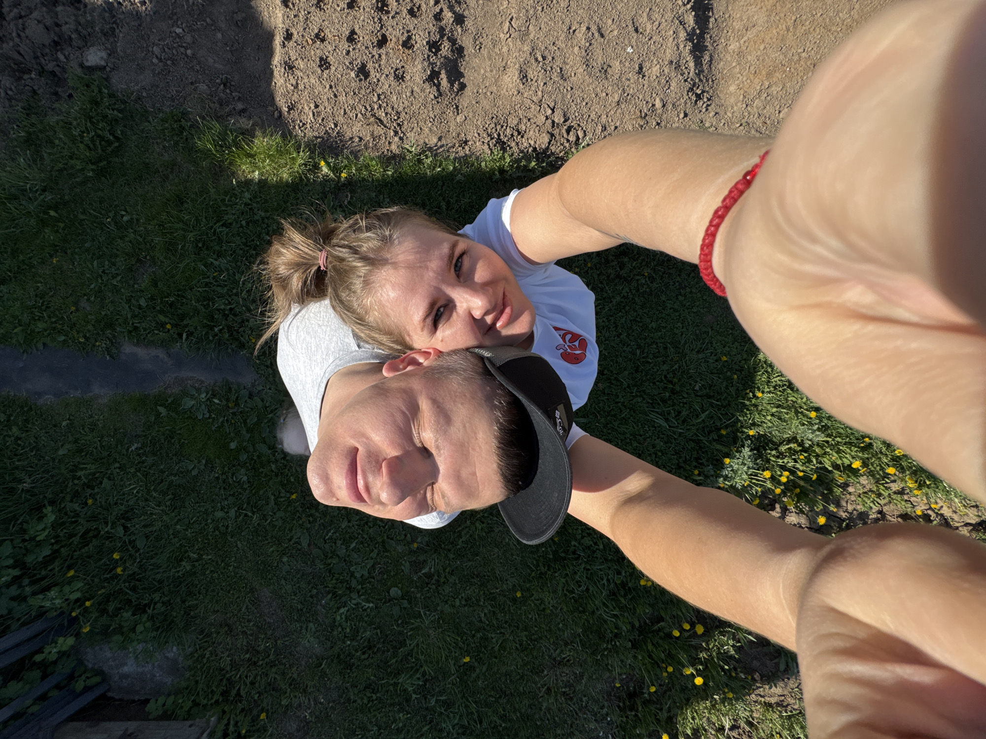 A high-angle shot shows a man and a woman, presumably taking a selfie. The woman is behind the man and to the right in the frame. The man is wearing a black baseball cap. The woman has her hair up in a ponytail. Both are wearing white t-shirts. The location appears to be outdoors in a grassy area next to a dirt patch. The photograph does not provide information about the location within Фоки, Russia.