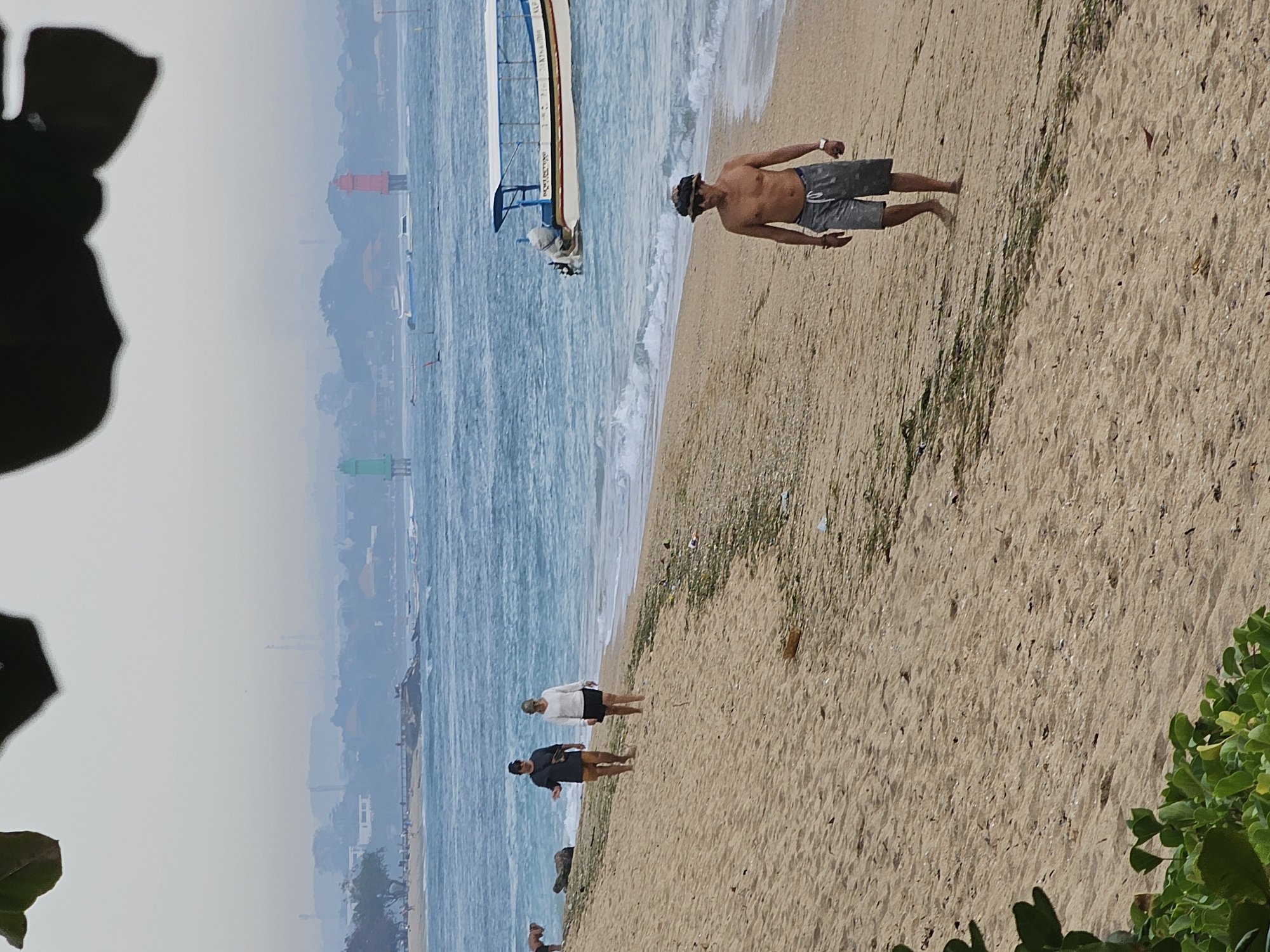 The image depicts a cloudy day on a beach in Denpasar, Indonesia. In the foreground, a shirtless man wearing grey shorts and a hat walks along the sandy shore. Behind him, two people, a man and a woman, walk together on the beach. In the distance, a boat with a blue canopy is anchored in the ocean, and two lighthouses, one red and one green, are visible. The city skyline is shrouded in haze.