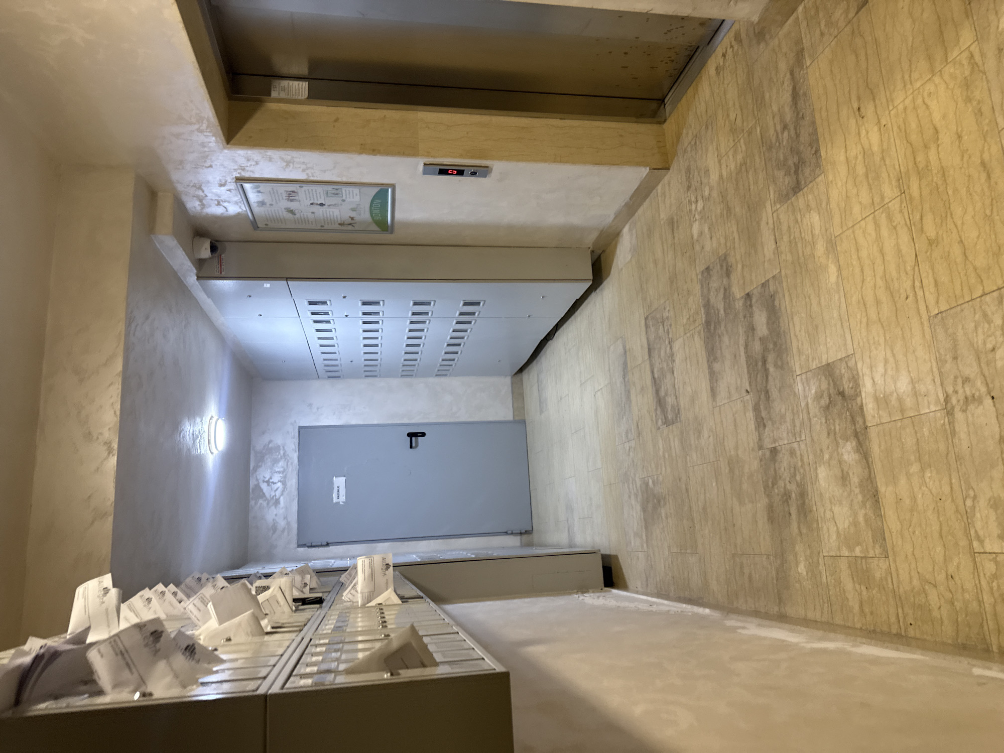The image depicts a hallway in Boreti, Montenegro, featuring mailboxes on the left, a grey door with a black handle in the middle, and an elevator on the right. The hallway is lit by an overhead light and has tiled flooring. A security camera is visible mounted on the wall above the mailboxes. A poster is displayed on the wall next to the elevator. The scene appears to be empty of people, with mail visible in the mailboxes and papers on the poster.
