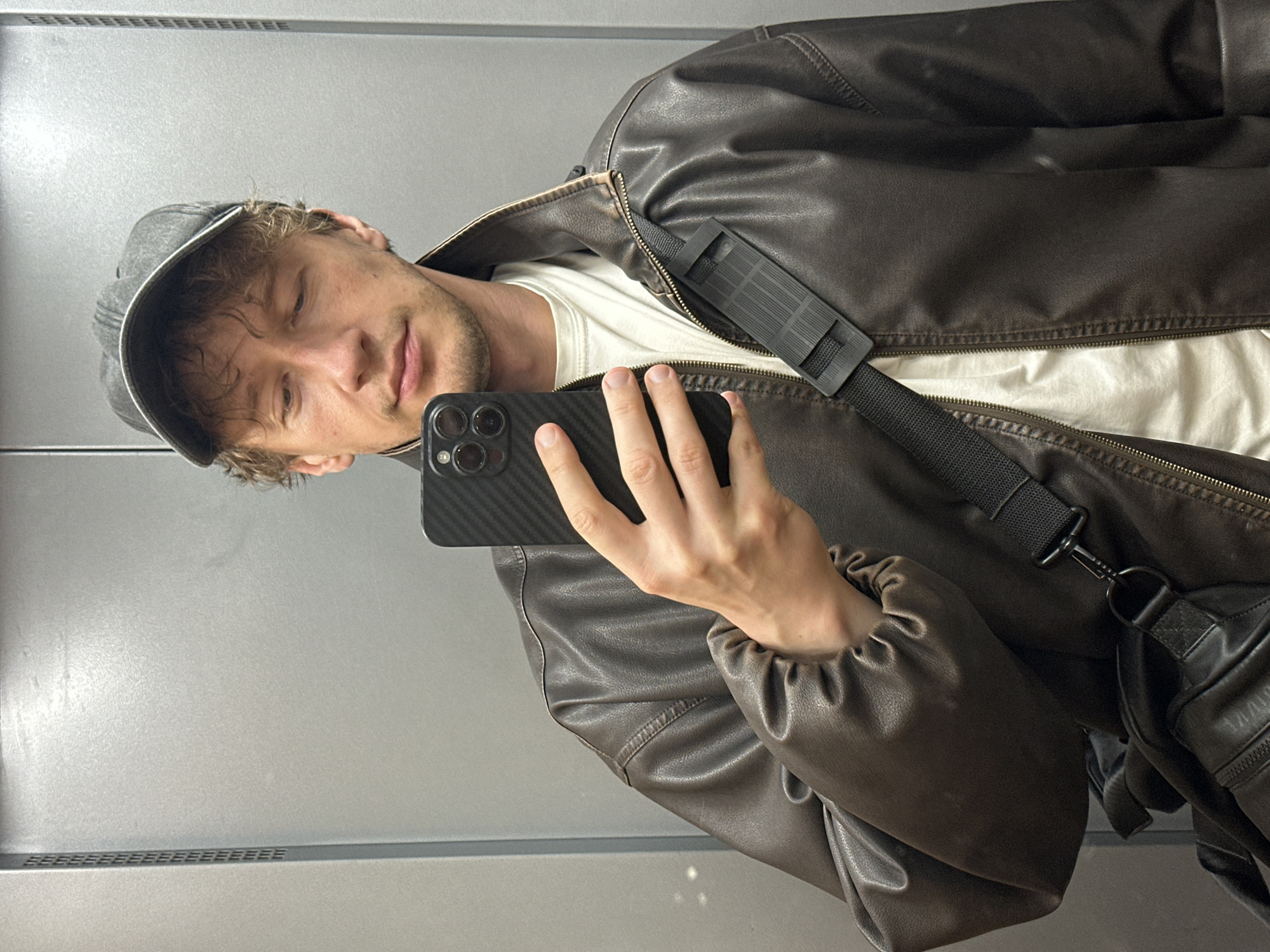 A man wearing a baseball cap and a brown leather jacket takes a selfie in an elevator. He is holding a smartphone in his right hand, and a black bag is slung across his body. The elevator walls are gray. The image was taken in Murino, Russia.