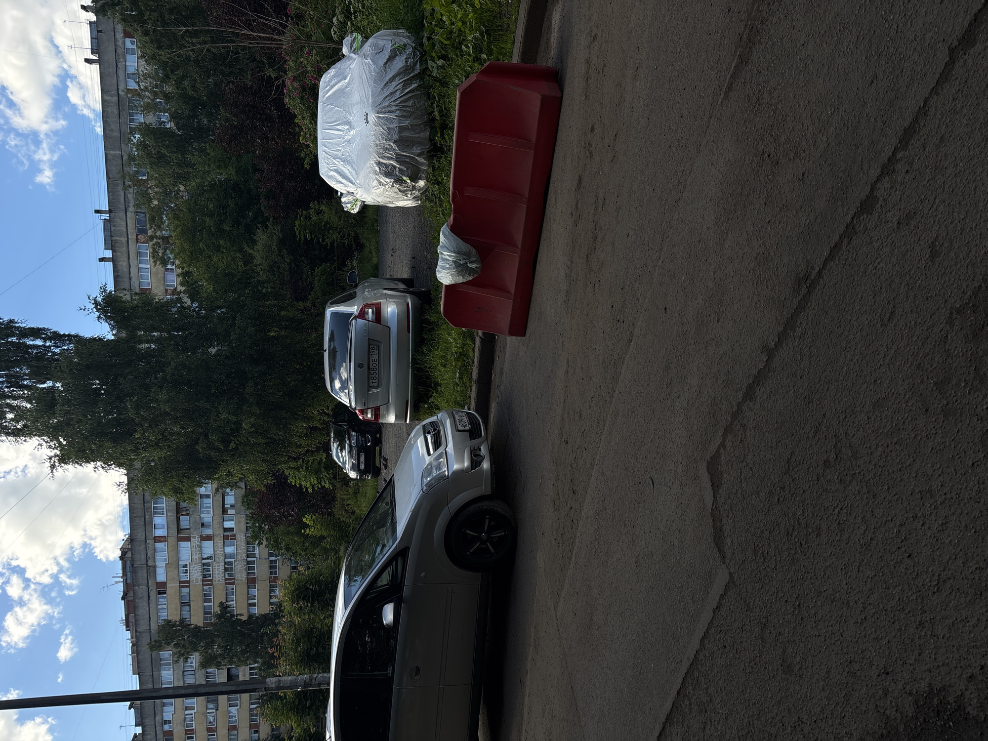 The image depicts an outdoor scene, likely in Saint Petersburg, Russia, with a focus on an urban residential area. In the foreground, a red plastic barrier is positioned on a paved surface, with a white plastic bag draped over its top. To the left, a silver minivan is parked. Behind the minivan, a silver sedan is visible. Further back, another car, a black SUV, is parked. In the background, a multi-story apartment building dominates the skyline. Tall green trees and foliage surround the parked vehicles and the building. The sky is partly cloudy, with blue sky visible between the white clouds. The overall impression is that of a typical urban environment, possibly a courtyard or parking area adjacent to an apartment complex.