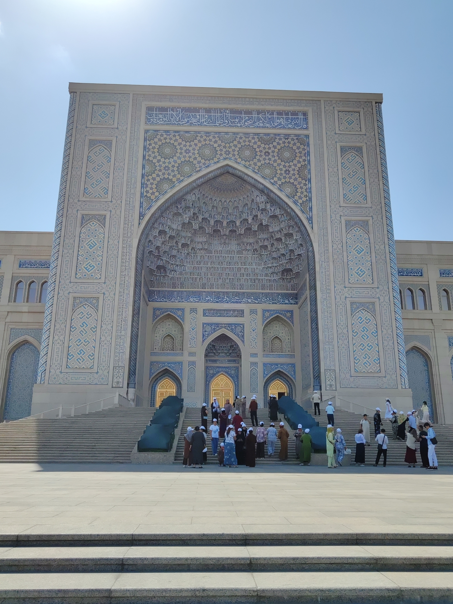 A wide, low-angle shot captures the grand entrance of the Hazrati Imam Mosque in Tashkent, Uzbekistan, on a clear, sunny day. The mosque's facade is adorned with intricate blue and white geometric patterns and Arabic calligraphy. A wide, shallow staircase leads up to the main entrance, with a group of people gathered on the steps and in front of the building. Most of the individuals appear to be tourists or worshippers, dressed in a mix of casual and traditional attire, with some wearing headscarves. Sunlight illuminates the scene, casting shadows on the stone pavement. The architecture is a blend of traditional Islamic design with modern construction elements.