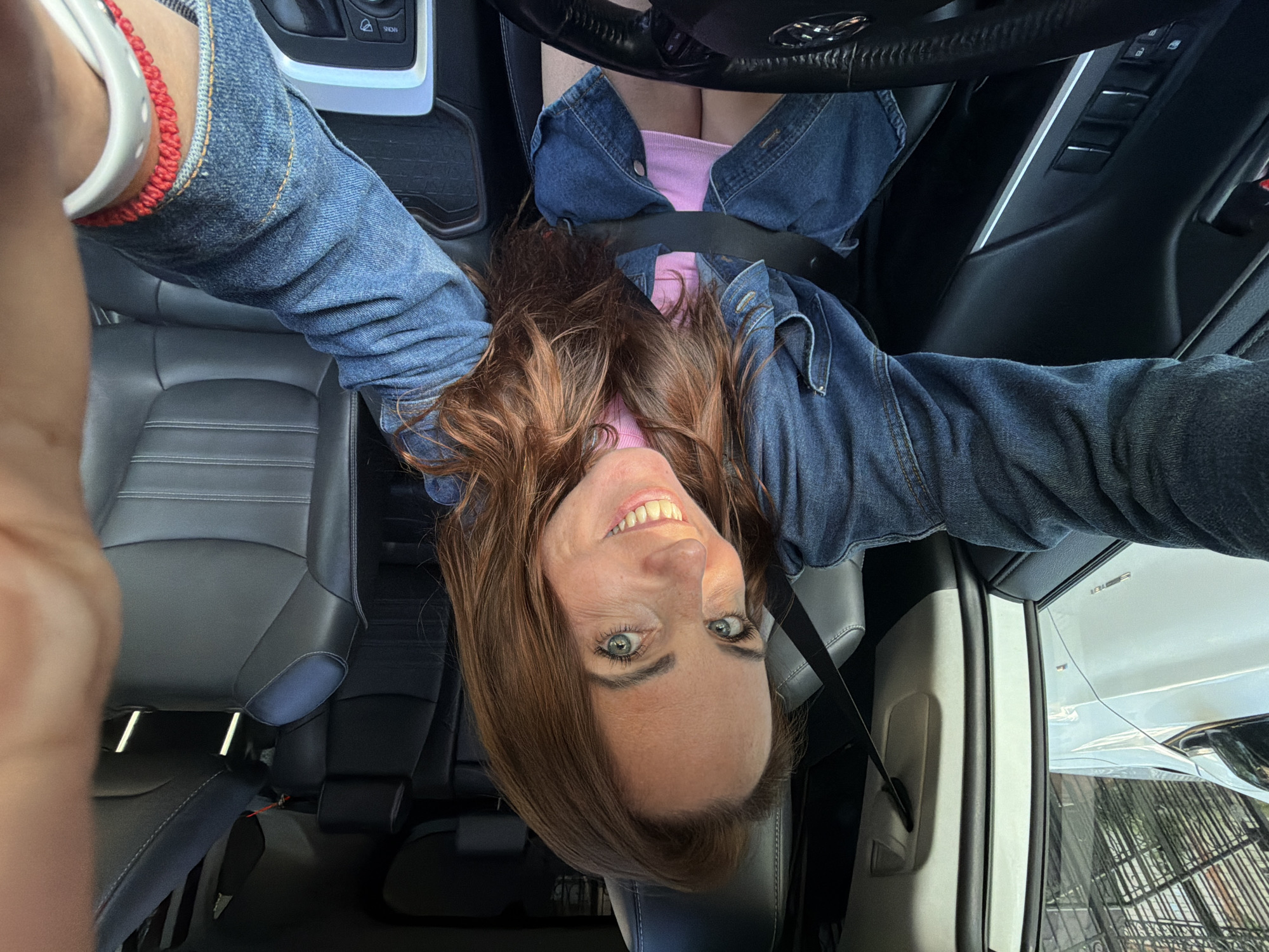 A woman with reddish-brown hair is taking a selfie inside a car. She is wearing a denim jacket over a pink top and has a seatbelt on. The car's interior is visible, with black leather seats and a steering wheel. Outside the car window, a white car and a metal fence can be seen. The scene is set in a city environment.