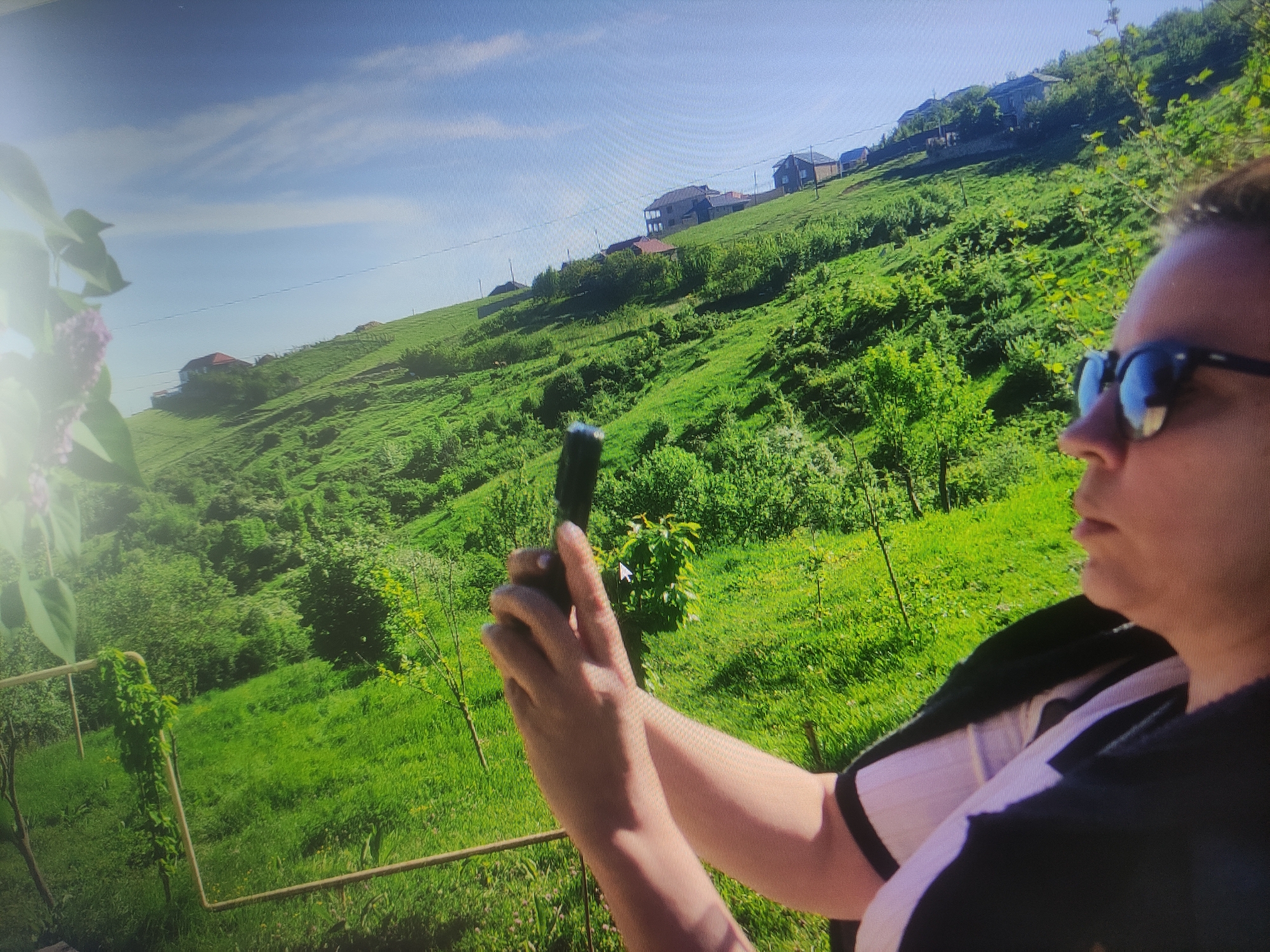The image captures a sunny day in a hilly, green landscape, possibly in the outskirts of Baku, Azerbaijan. The foreground is dominated by a woman with sunglasses, holding what appears to be a dark-colored object, possibly a phone or camera, in her hands. Her arms and shoulders are visible, and she is wearing a dark top with white accents. The background shows a verdant hillside dotted with houses, suggesting a residential area. The sky is blue with scattered clouds. The overall impression is of a bright, outdoor scene.