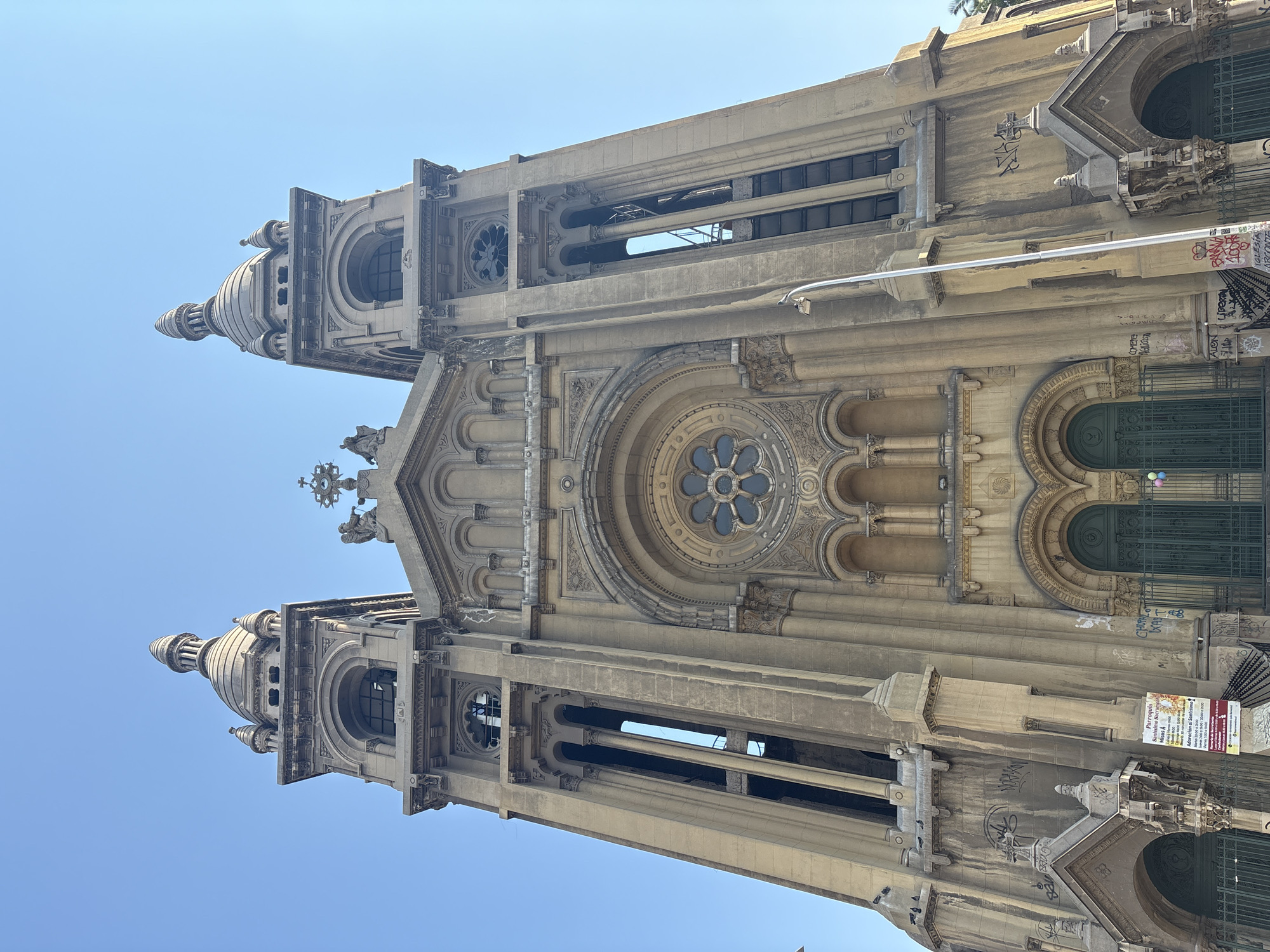 This is a low-angle, eye-level, full-shot photograph of the Parroquia Santísimo Sacramento in Santiago, Chile. The church, constructed of light brown stone, is bathed in sunlight under a clear blue sky. The facade features two prominent bell towers, each topped with a domed spire and smaller decorative turrets. Intricate architectural details, including arched windows, decorative carvings, and a rose window above the main entrance, are visible. The entrance consists of two large, dark green double doors, flanked by arched doorways. The building exhibits some signs of graffiti. A street lamp stands in front of the right tower. A sign on the left side of the facade displays information about the church. A flock of birds is perched on the left side of the building. The overall scene depicts the exterior of a historic religious building.