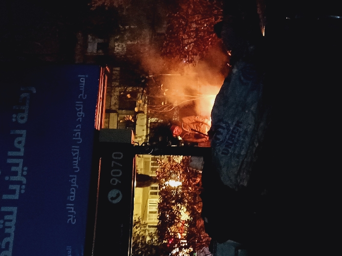 A nighttime scene shows a man near a fire or explosion in an urban area. A blue sign with Arabic text is visible on the left, advertising a "Journey to Malaysia." A telephone number, 9070, is also on the sign. The fire casts an orange glow illuminating the immediate surroundings, while the rest of the scene is shrouded in darkness. Trees and buildings are faintly visible in the background, hinting at a street or residential area.