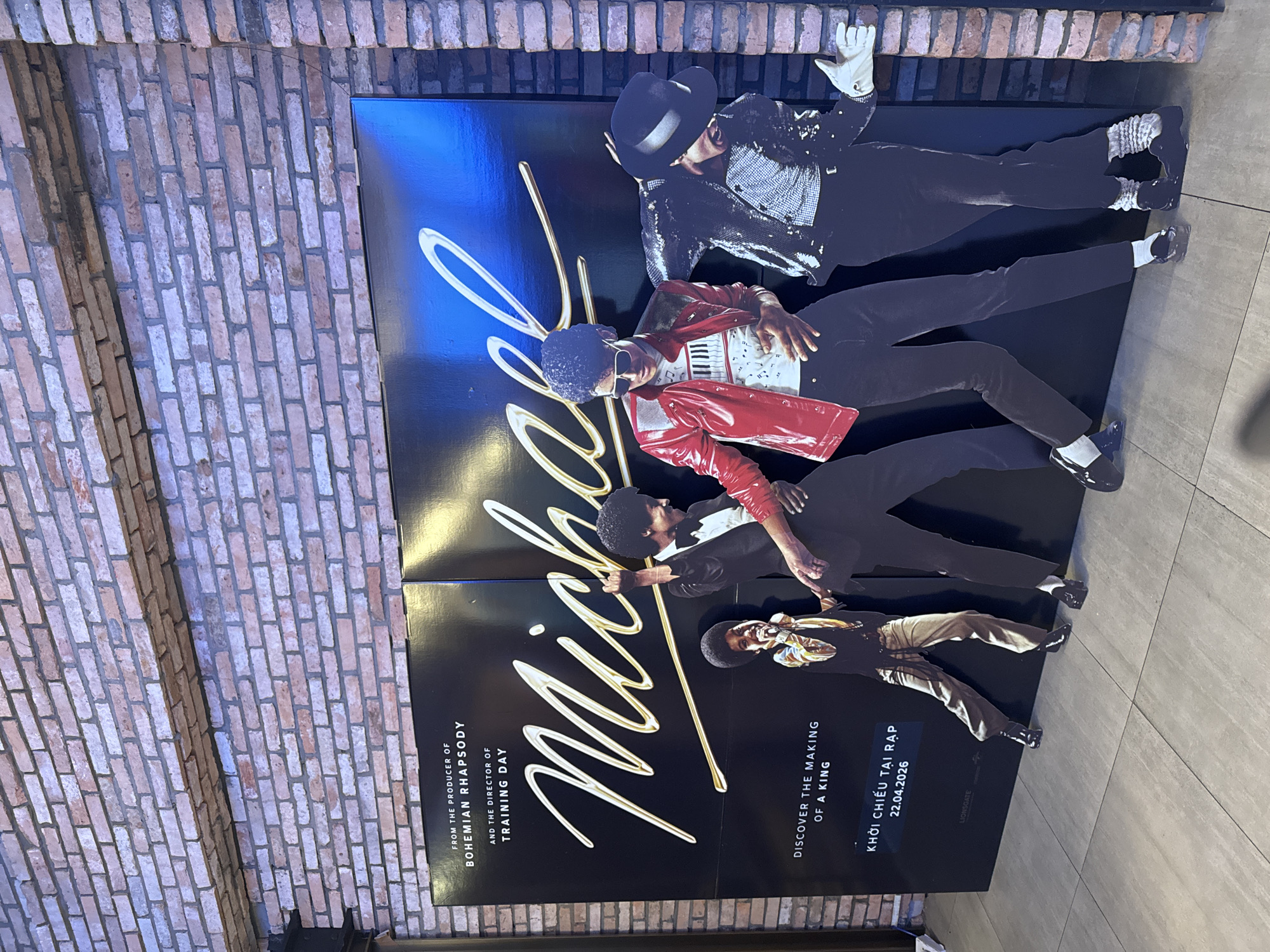 Here's a concise description of the image:
The image captures a promotional poster for the movie "Michael," a biographical film about Michael Jackson. The poster features five cutout images of Michael Jackson in various iconic outfits and poses, layered over a black background with "Michael" written in large, stylized gold lettering. The poster also includes text indicating the film is from the producer of "Bohemian Rhapsody" and the director of "Training Day." It states "Discover the Making of a King" and the release date in Vietnamese: "KHỞI CHIẾU TẠI RẠP 22.04.2026." The poster is positioned near a brick wall. The setting appears to be inside near a marble looking floor. No persons can be identified.