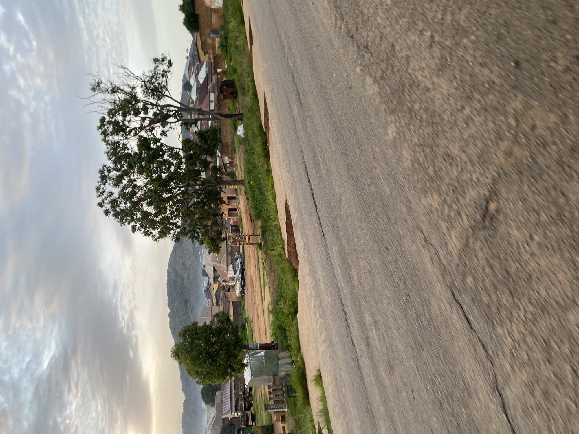 The image depicts a scene in Gwong, Nigeria, under a cloudy sky. In the foreground, a cracked asphalt road stretches diagonally across the frame. To the left, beyond the road, is a dirt area with various objects. These include a silver corrugated metal structure, several vertical stone-like pillars, and what appears to be part of a truck with a flatbed. Further back, a small group of buildings with red and brown roofs are visible, nestled against a backdrop of lush green hills and trees. Several cars are parked in the open area between the road and the buildings. Two prominent trees, one with dense foliage and another with sparser branches, stand out in the middle ground. The overall impression is one of a semi-rural or peri-urban environment.