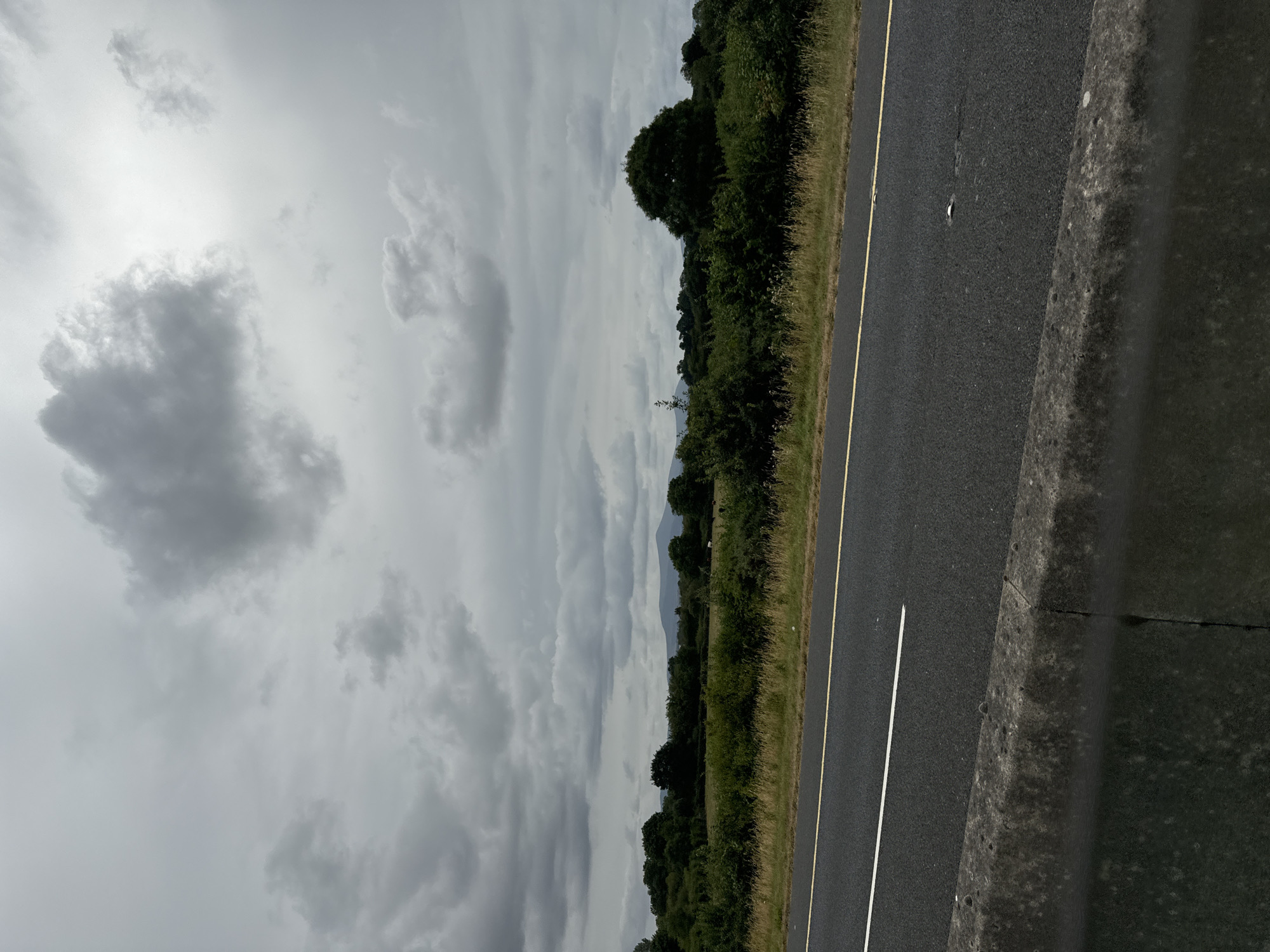 The image shows a grey, overcast sky with dark clouds. Below the sky is a landscape with rolling hills and trees. In the foreground, there is a road with a white line and a yellow line, and a concrete barrier. The scene appears to be in a rural area, possibly in Ireland, given the mention of Garryduff Lane. There are no people or animals visible in the image. The overall mood is somber and perhaps a little dramatic due to the stormy sky.