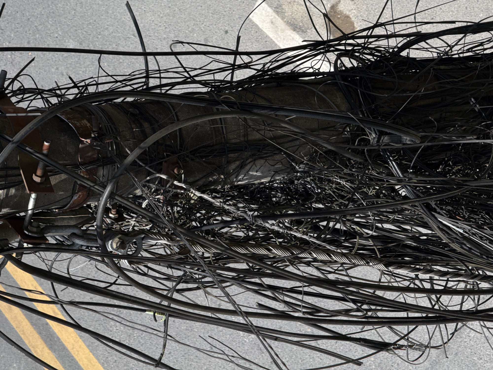 This close-up, eye-level shot captures a tangle of black electrical and communication wires wrapped haphazardly around a concrete utility pole. The pole itself is rough and textured, with visible metal brackets and rusty bolts securing some of the wiring. The wires vary in thickness and composition, some appearing to be insulated cables, while others are braided metallic strands, possibly structural support wires. They cascade down the pole and spill out to the sides, obscuring much of its surface. In the background, a grey asphalt surface is visible, marked with two parallel bright yellow road lines, indicating a street or roadway. The lighting suggests a daytime scene, with shadows and highlights defining the complex network of wires. The overall impression is one of urban clutter and infrastructure inefficiency, a common sight in many cities, potentially including Dalat, Vietnam, where such dense and unorganized wiring is prevalent in certain areas. There are no people or animals visible in the image, and no specific actions are being depicted beyond the static presence of the tangled wires.