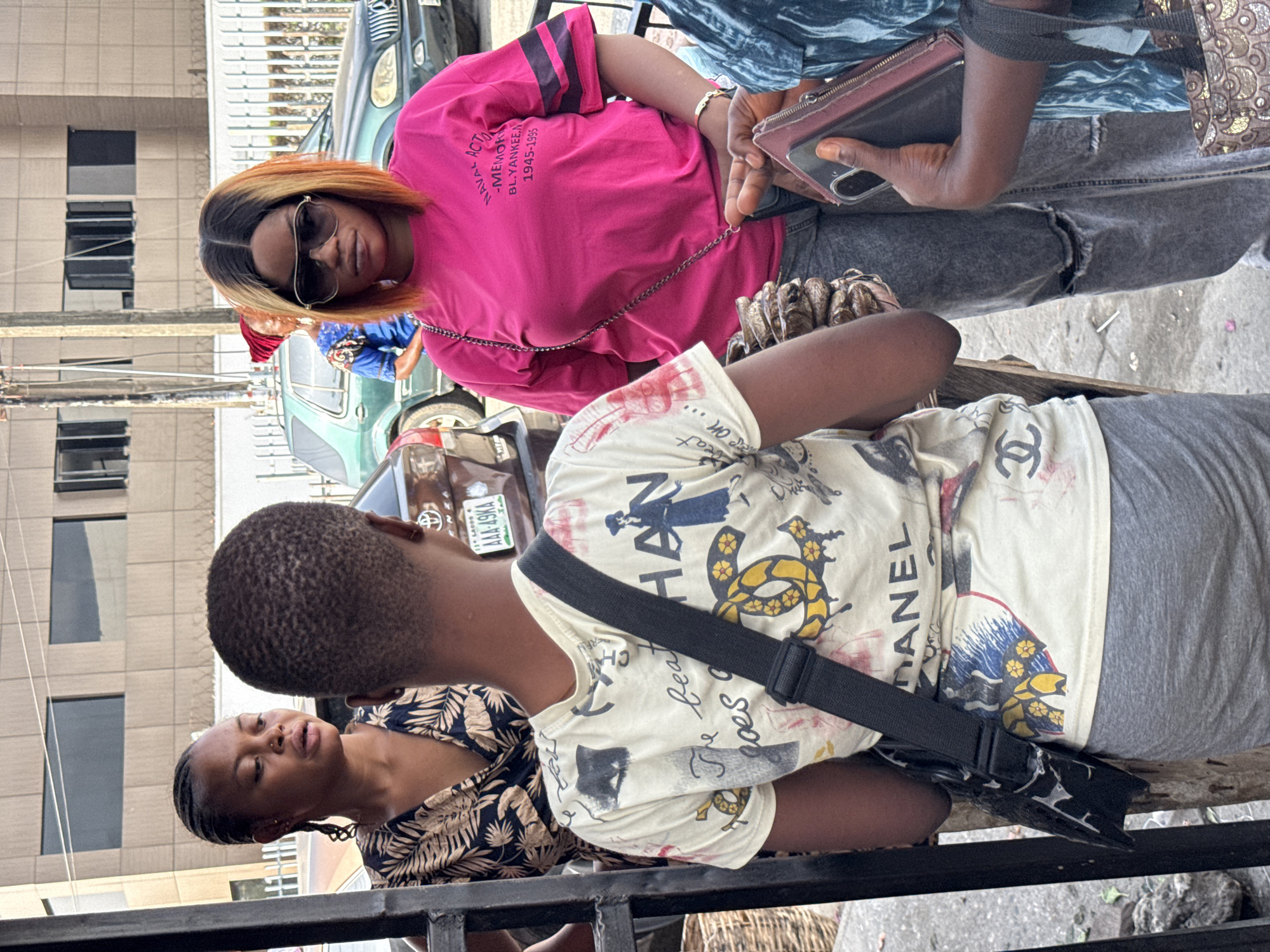 The image depicts a street scene, possibly in Ado, Nigeria. The focus is on a group of people standing together. A woman in a pink shirt with the inscription "ACT NAVAL - MEMOR BL.YANKEE.A 1945-1995" is seen wearing sunglasses. Another person is wearing a cream-colored t-shirt with dark prints and a black shoulder bag. The shirt has the printed word "CHANEL". Another woman in a printed top is also visible. A car can be seen in the background with a license plate marked "LAGOS AAA-49KA". A building is also visible in the background. The people appear to be engaged in conversation or waiting.