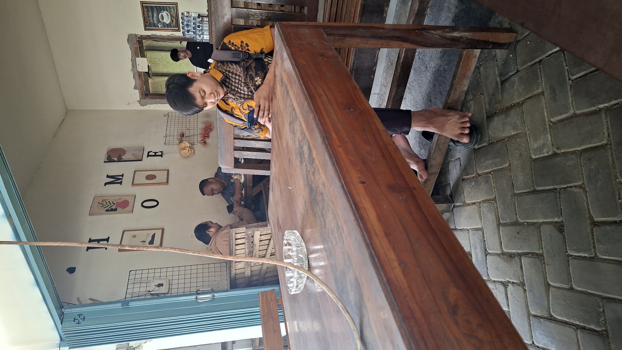 A medium shot captures a man sitting at a wooden table in a cafe in Ngimbang, Indonesia. He is looking down at his smartphone, which he holds in his hands. He wears a yellow shirt with a patterned design and dark pants. His bare feet are visible, resting on the paved floor, with one foot in a black sandal. Behind him, two other men are seated at tables, also appearing to be engaged with their phones. The cafe's interior features white walls decorated with framed art and the word "HOME" spelled out in large letters. Wooden furniture, including tables and benches, is present throughout the space. A glass ashtray sits on the table in the foreground. The environment suggests a casual, relaxed atmosphere.