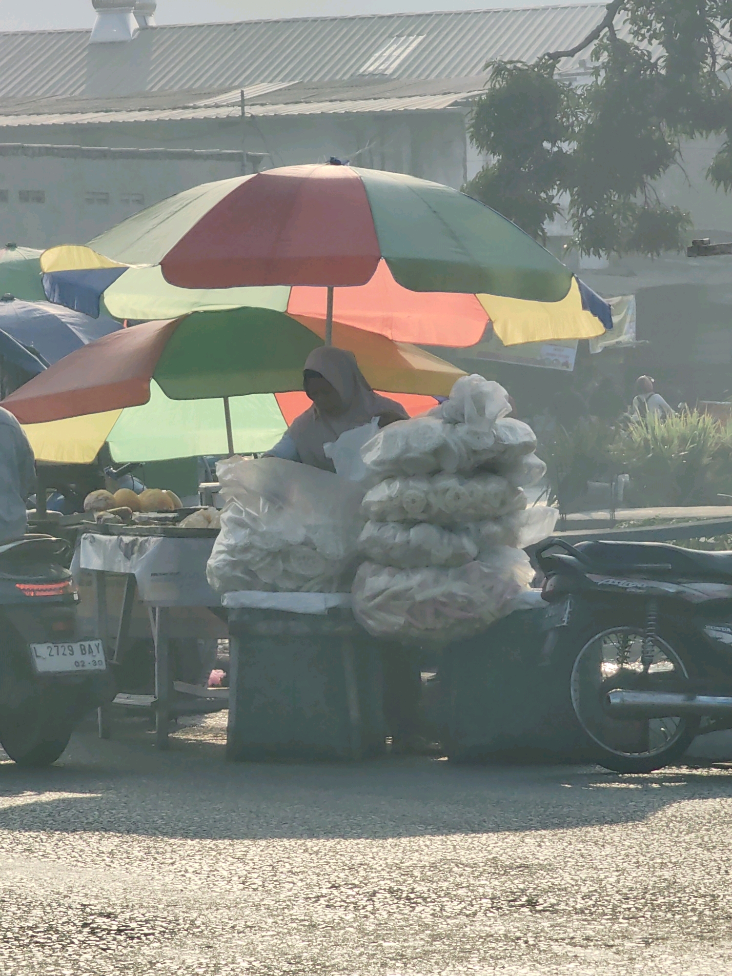 The image captures a street scene in Keboansikep, Indonesia. A vendor, wearing a hijab and a light-colored top, is seen behind a cart laden with goods. The cart displays several yellow, round items, possibly food, and is covered with white plastic bags filled with what appear to be snacks or puffed crackers. Over the cart, multiple colorful umbrellas (red, green, yellow, and blue) provide shade. In the foreground, a motorcycle with the license plate "L 2729 BAY" is parked on the right side. Another motorcycle is partially visible on the left. The background shows buildings with corrugated roofs and some greenery, suggesting an outdoor market or roadside stall environment. The lighting indicates it is daytime, and a slight haze is present, characteristic of the local atmosphere.