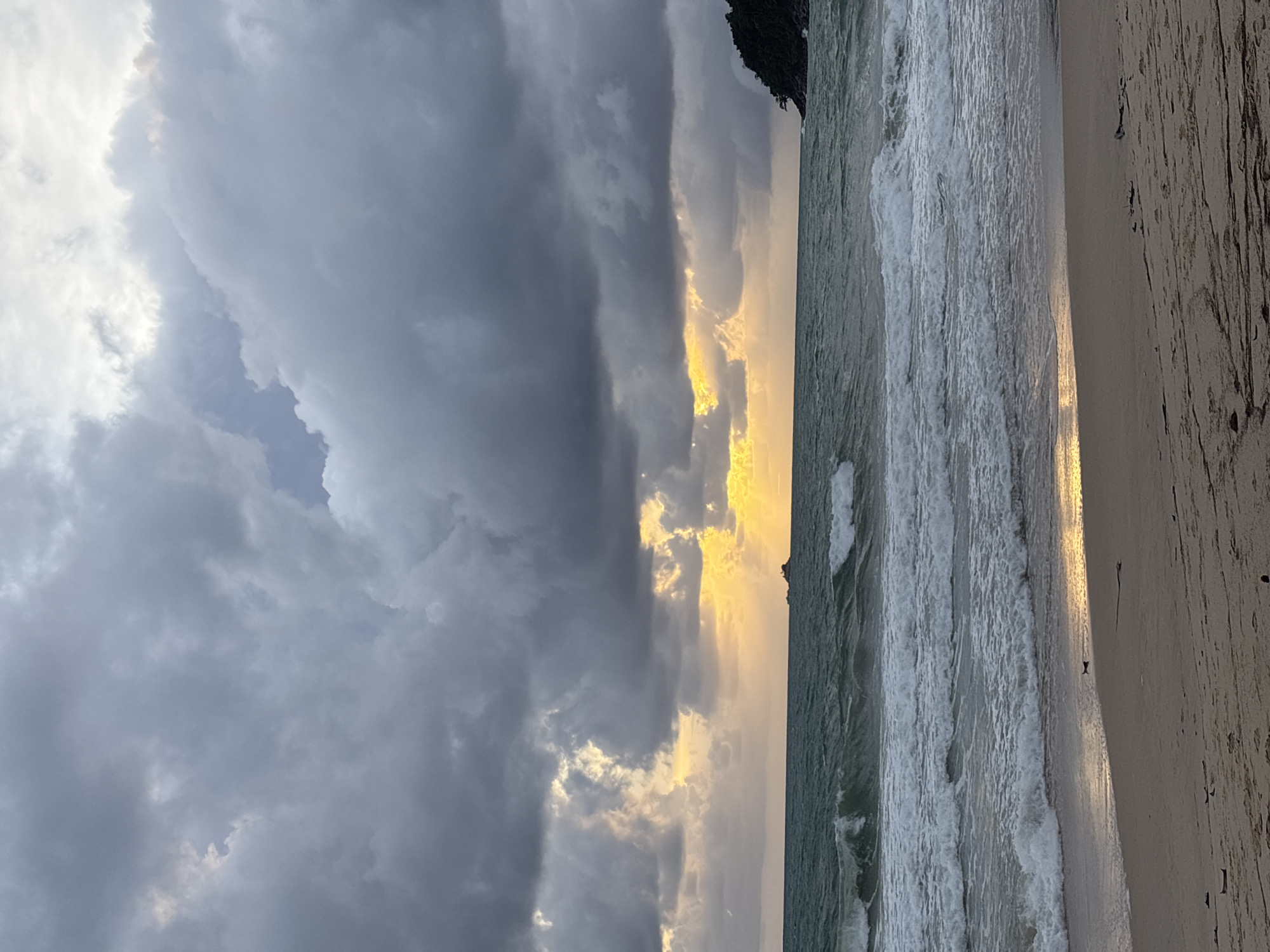 The image depicts a beach scene in Choeng Thale, Thailand. The sky is filled with dramatic clouds, ranging from bright white to dark gray, with a streak of golden sunlight breaking through. The ocean has choppy waves and the beach is sandy with some debris or textures. The photo is taken with a slight sideways angle.