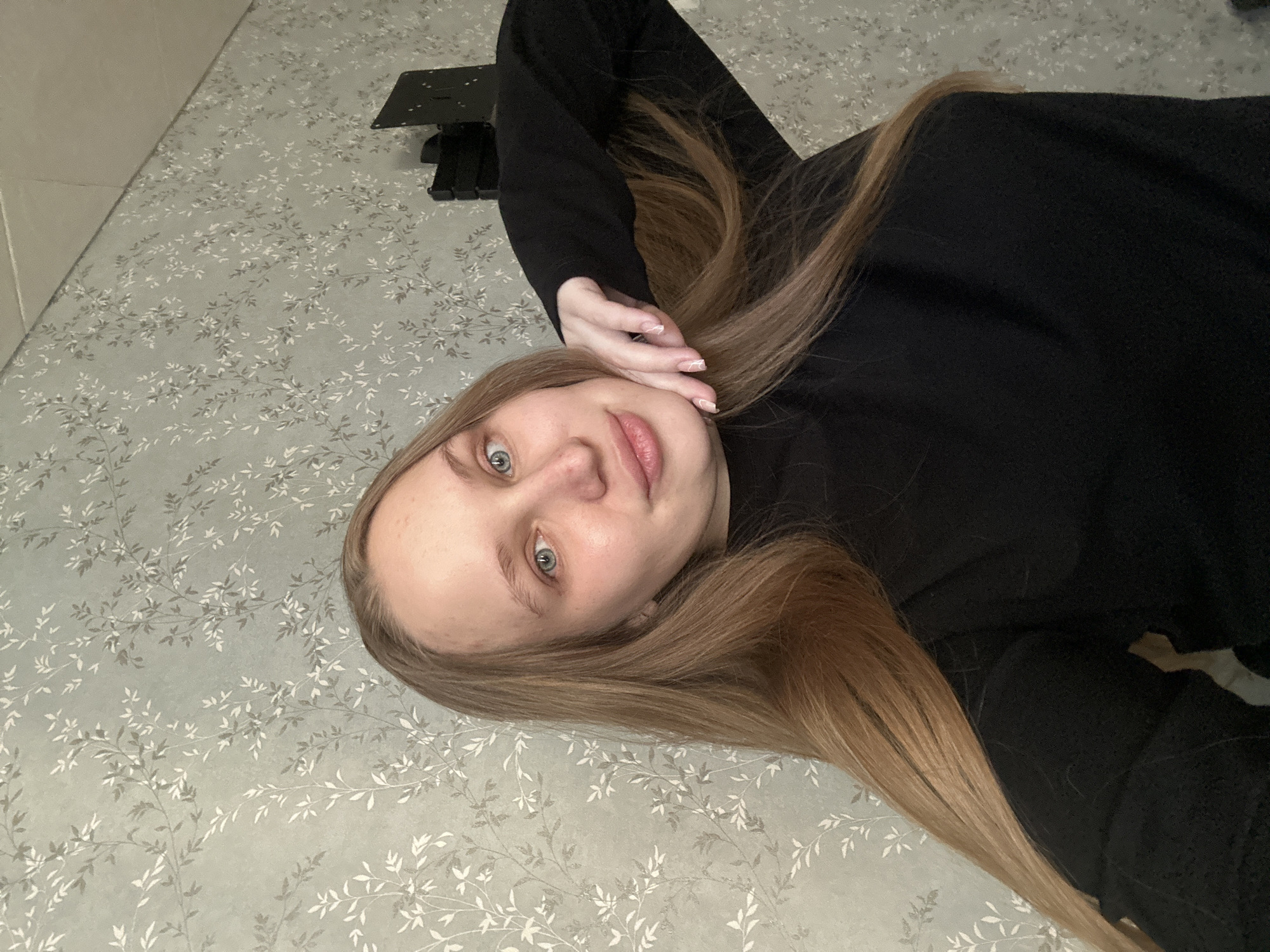 A young woman is lying on a bed with a floral-patterned bedspread. She is wearing a black sweater. Her long, light brown hair is spread out on the bed. Her right hand is near her face. Behind her head is a light-colored wall. In the background, there is a black object. The city location (Chaikovsky, Russia) is not determinable from the visual content of the image.