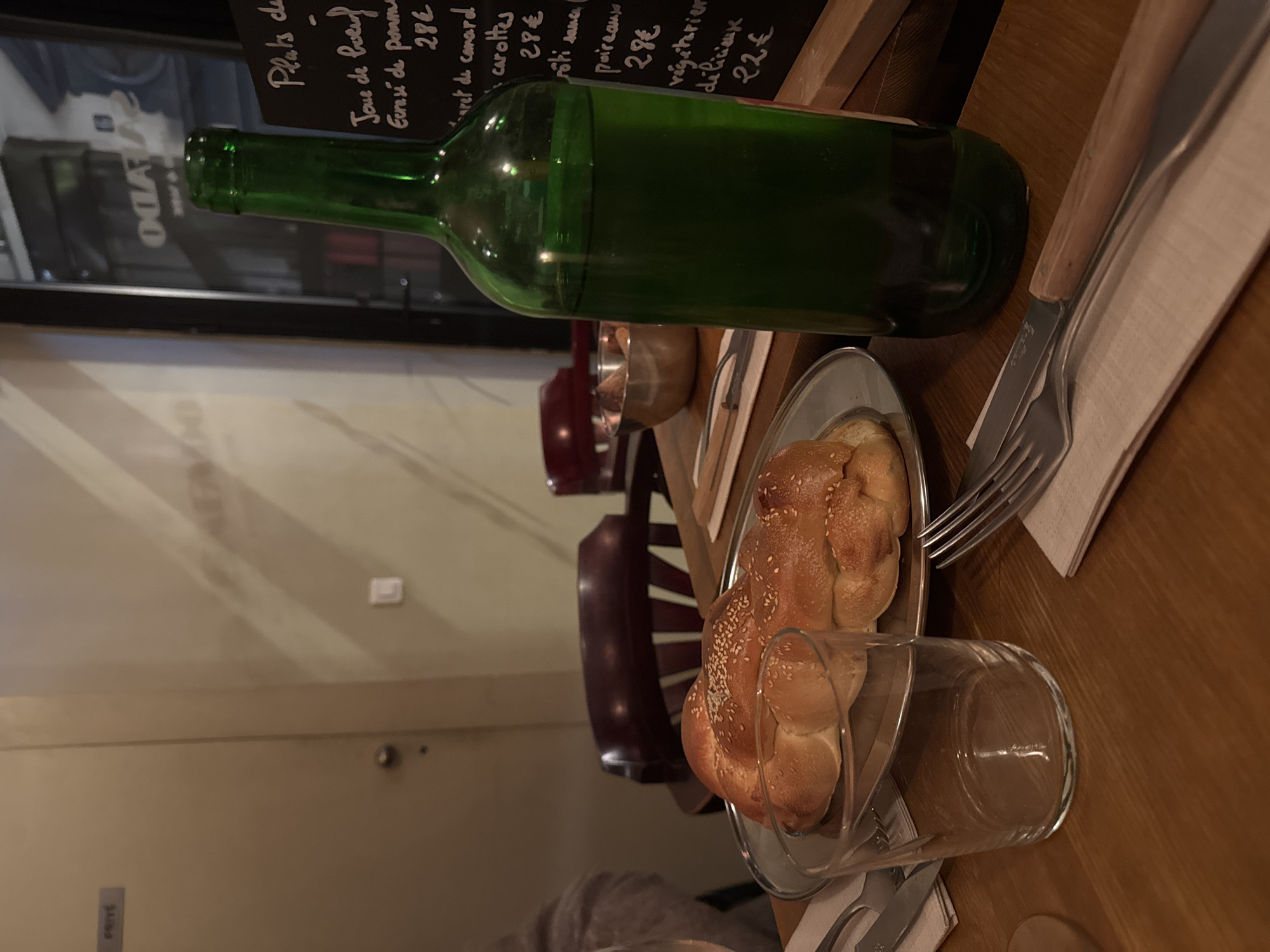 The image depicts an indoor restaurant setting at a wooden table in Paris, France. In the immediate foreground, an empty clear glass tumbler rests on the table, to the right of which is a folded light-colored napkin holding a silver fork and knife with light-colored wooden handles. Centrally positioned on the table is a silver-colored oval platter containing two braided loaves of bread topped with sesame seeds. To the right of the bread platter, a dark green glass bottle, approximately half-full of a green liquid, stands upright. In the mid-ground, a stainless steel bowl containing an indeterminate food item is visible, along with parts of a dark red upholstered chair. A person's arm, covered in light grey fabric, is partially visible on the far left. The background features a light-colored wall with a closed door marked with a rectangular "PRIVÉ" (Private) sign and a dark door handle. To the right, a dark-framed glass partition or door reflects parts of the interior and displays reversed text, appearing as "OGIA." Adjacent to this is a dark chalkboard menu, handwritten in white, listing French dishes such as "Joue de boeuf" (beef cheek), "Écrasé de pomme" (crushed potato), "Confit de canard" (duck confit), "carottes" (carrots), "poireaux" (leeks), and "végétarien" (vegetarian) options, with prices ranging from 22€ to 28€. The overall lighting is dim, characteristic of an evening dining ambiance.
