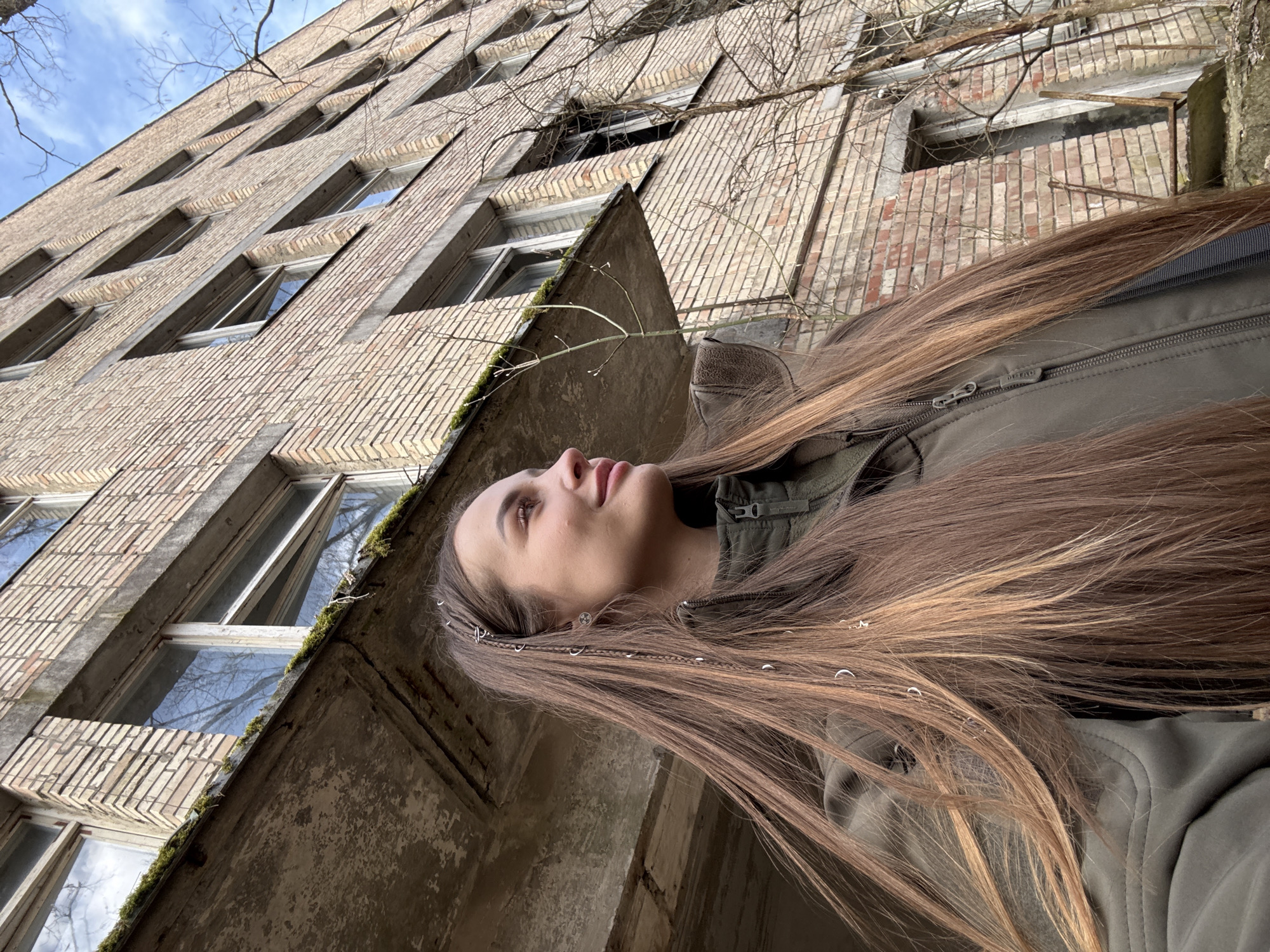 A young woman with long brown hair stands outdoors in Prypiat, Ukraine, looking up at a tall, dilapidated brick building. The building is multiple stories high, with many windows, some of which are open or broken. There are patches of moss growing on the exterior of the building. The woman wears a green jacket. The sky is partly cloudy.