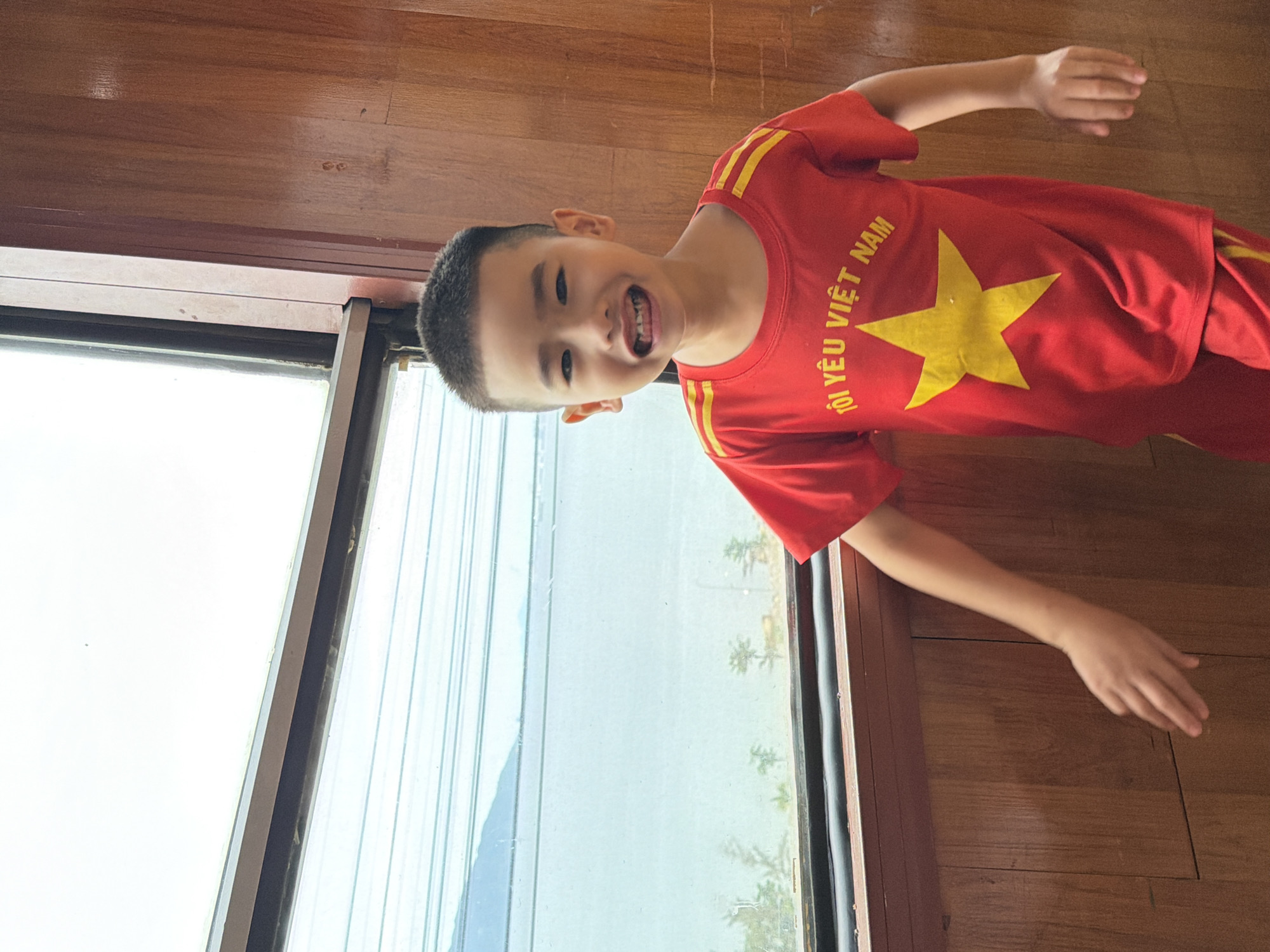 A young boy with short dark hair and an open mouth smiles broadly while standing indoors next to a large window. He is wearing a red t-shirt with a yellow star and the words "TÔI YÊU VIỆT NAM" printed on it, and red shorts with yellow stripes. The window shows a hazy view of the ocean and distant mountains. The boy's right hand is extended towards a wooden wall. The image appears to have been taken in Đà Nẵng, Vietnam, possibly from a room overlooking the coast.